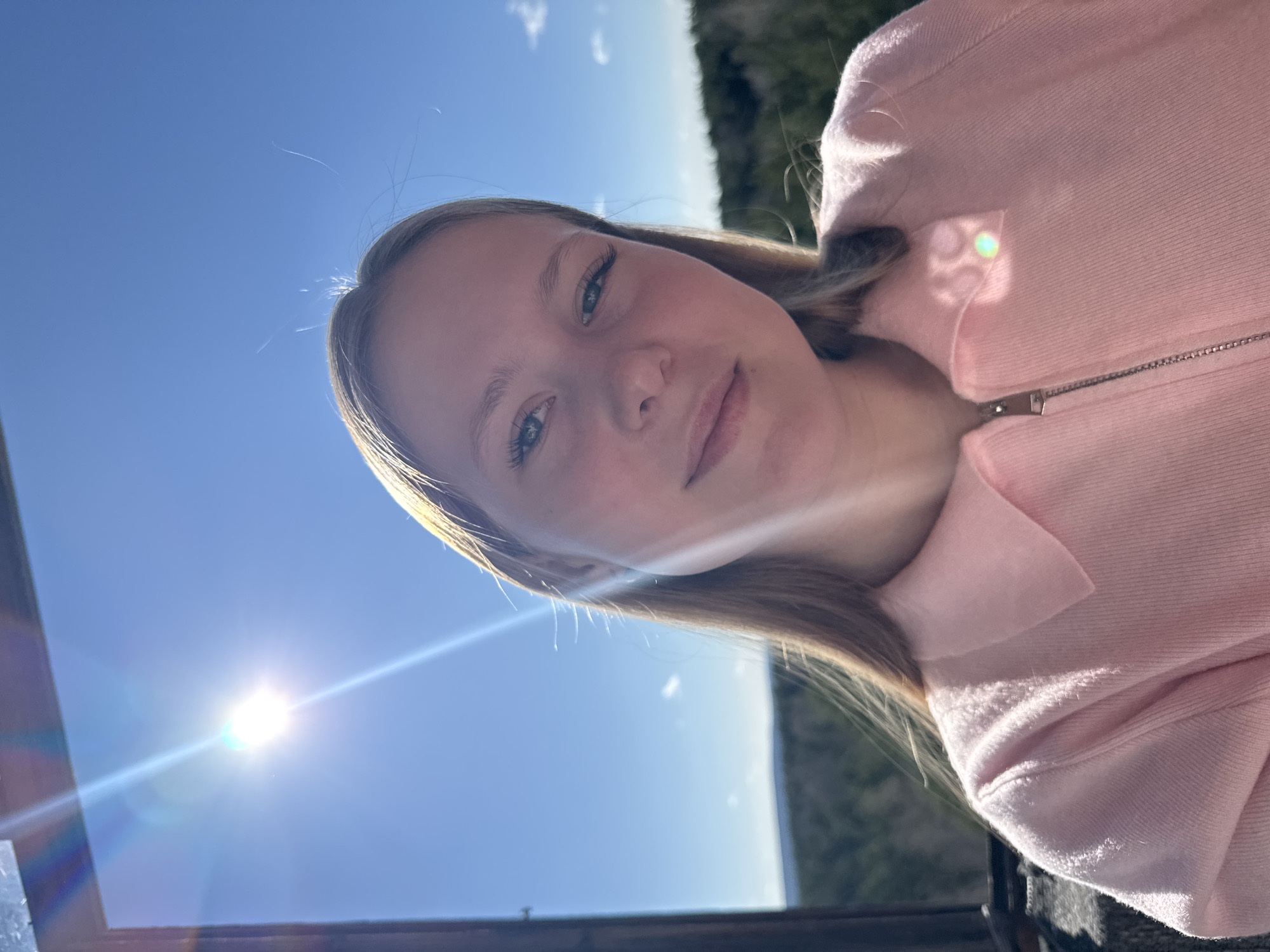 A selfie of a young woman wearing a pink jacket is angled upward against a bright, blue sky with a bright sun visible in the lower left. The background consists of a distant, forested area. A portion of a structure is also visible in the lower left. The image was most likely taken outside on a sunny day. Information about the location in the city of Chaikovsky, Russia cannot be inferred from the image.
