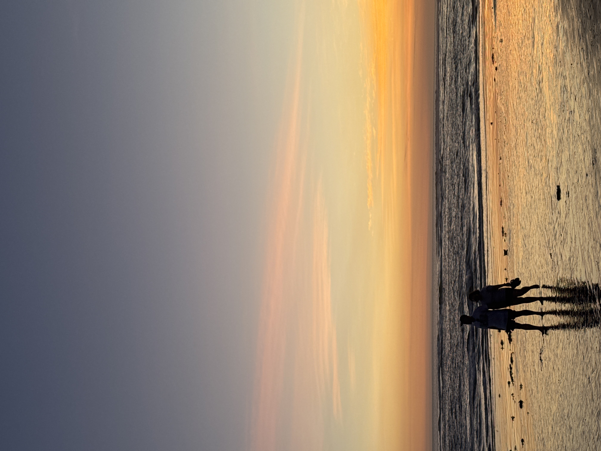 This image depicts a serene beach scene at sunset, with the sky transitioning from a soft gray to warm hues of orange and yellow. The sun is low on the horizon, casting a golden glow across the wet sand and the gentle waves. In the foreground, silhouetted against the vibrant sky, are two individuals walking along the shoreline. Their forms are indistinct, but they appear to be enjoying a peaceful stroll. Their shadows stretch long across the sand, indicating the low angle of the sun. The wet sand reflects the colors of the sky, creating a shimmering effect. The overall mood of the image is tranquil and contemplative, capturing the beauty of a sunset on a tropical beach. The location is North Kuta, Indonesia, a place known for its stunning coastal landscapes and breathtaking sunsets.