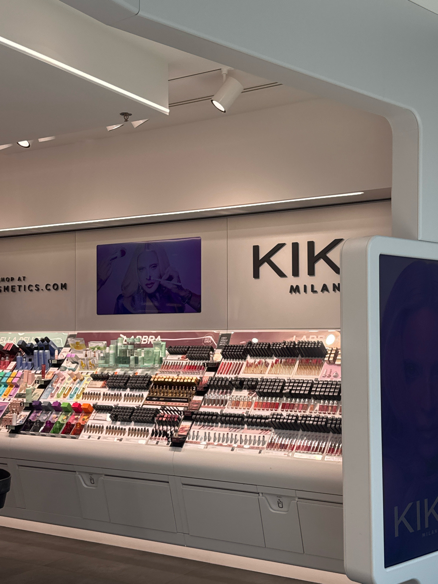 The image shows a brightly lit KIKO Milano cosmetics store, likely located in Carini, Italy, given the context. The store features a wide array of makeup products displayed on tiered shelves. Prominently displayed on the wall is the KIKO Milano logo, with the word "KIK" in large black lettering and "MILAN" in smaller lettering beneath it. To the left of the logo, a digital screen shows a close-up of a blonde woman applying makeup. The shelves are filled with various cosmetic items, including lipsticks, glosses, and other beauty products, arranged in an organized manner. In the foreground, a display case with drawers is visible. The lighting is modern and sleek, with recessed ceiling lights and strip lighting along the shelves. The overall impression is of a clean, well-stocked, and modern retail environment.