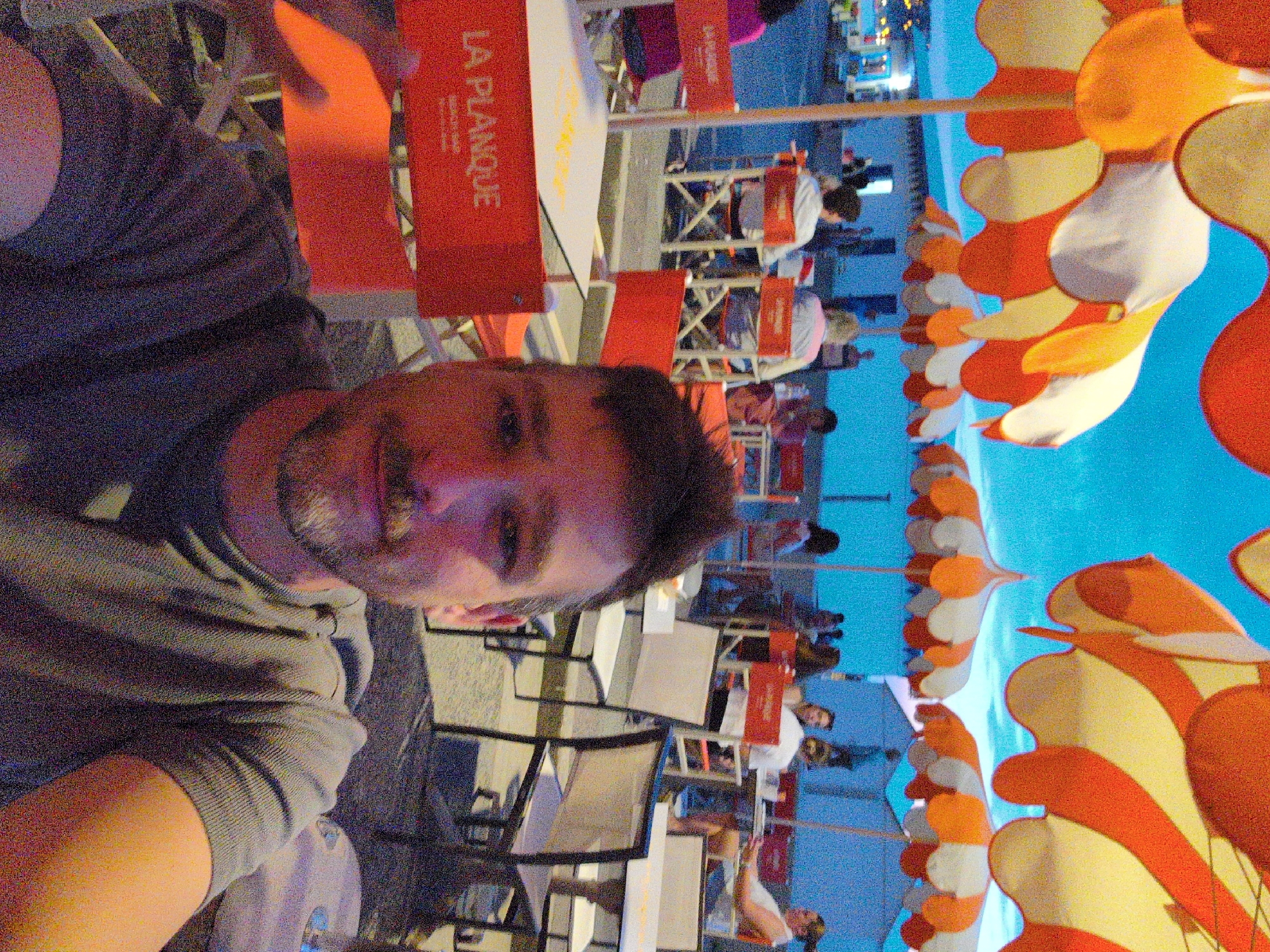 A man with a beard and mustache takes a selfie in a restaurant with a blue sky and orange and white umbrellas. He is wearing a gray t-shirt. In the background, there are tables and chairs, other people, and buildings. The restaurant is located in Mers-les-Bains, France.