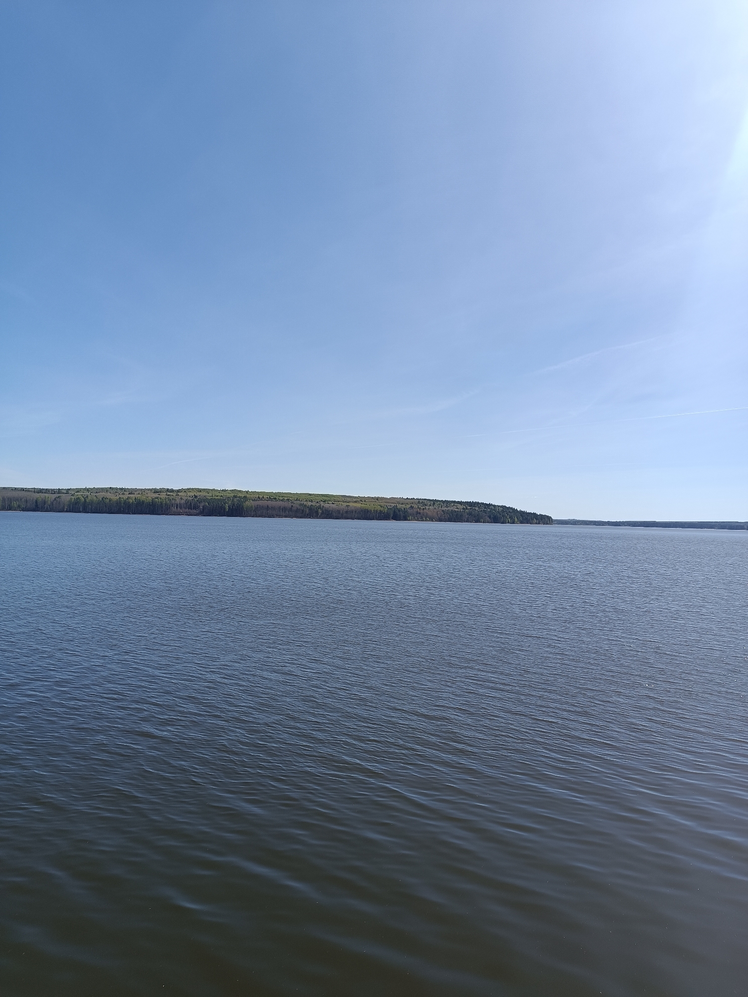 The image shows a vast expanse of water under a clear, light blue sky. A long, low landmass is visible in the distance, covered in trees. The water is dark and subtly rippled, reflecting the sky. The overall scene depicts a calm and open natural environment, likely a lake or large body of water. The location is the city of Chaikovsky, Russia.