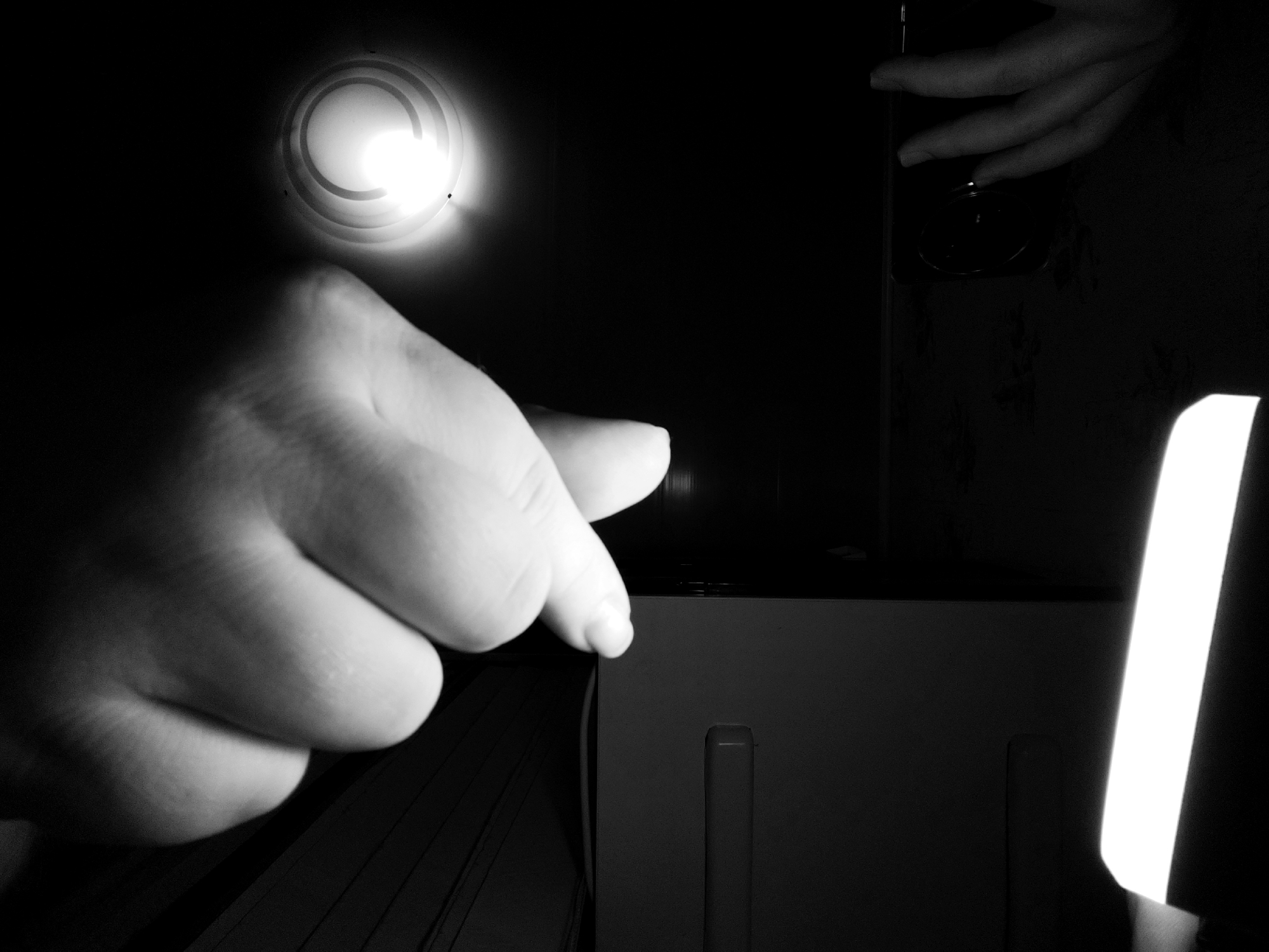 A close-up, black and white, low-angle shot focuses on a person's left hand in the foreground, with the index finger extended and pointing downwards towards the right edge of the frame. The hand is illuminated from above and to the left by a bright, circular light source, casting shadows and highlighting the texture of the skin. In the background, to the right, another hand is visible, its fingers curved and reaching downwards towards a dark, rectangular object, possibly a device. The environment is dimly lit, with a bright, vertical light source on the far right of the frame. The overall scene is abstract and suggestive, with no clear indication of its location within Fergana City, Uzbekistan.