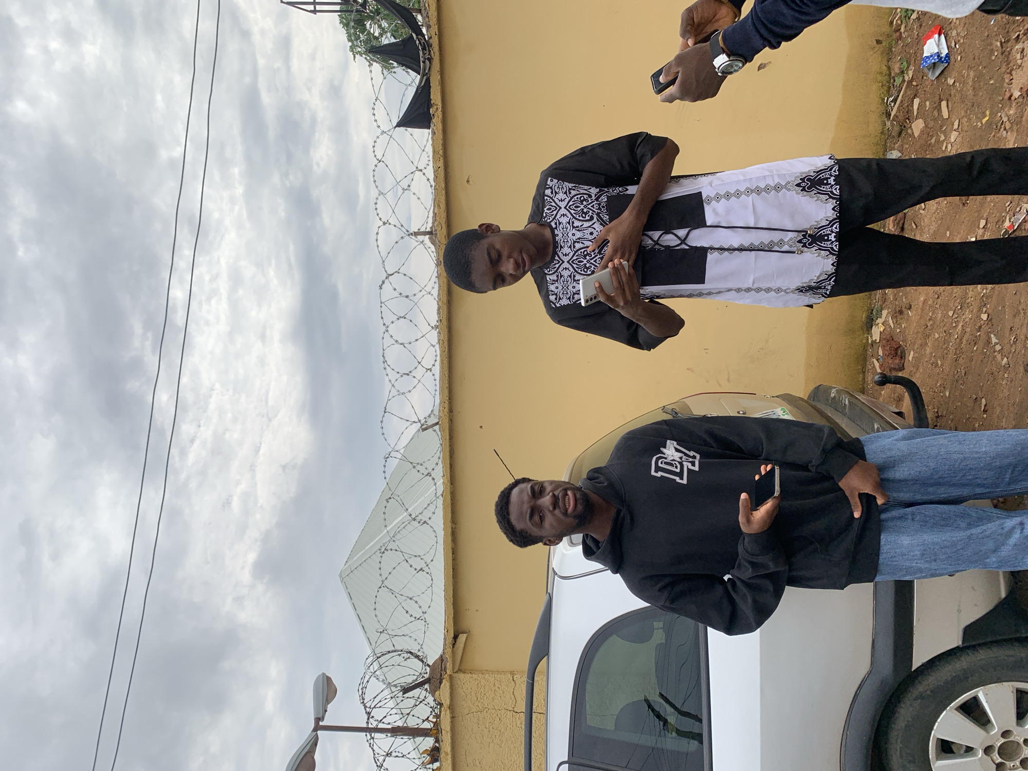 The scene is an outdoor setting in Jos, Nigeria, under an overcast sky. Three individuals are present. To the left, a young Black male with a short beard wears a black hoodie with a white "DY" logo and light blue baggy jeans. He holds a black smartphone in his left hand, partially tucked into his pocket, and looks towards the right with a slightly open mouth. Adjacent to a silver station wagon, which is partially visible on the left, an unpaved reddish-brown dirt ground with small debris covers the foreground.
In the center-right, a young Black male wears a black and white patterned long-sleeved tunic and black trousers. He holds a white smartphone with both hands at chest level, looking down at its screen. His left hand rests over his chest.
To the far right, only the hands and forearms of a third person are visible, wearing a dark long-sleeved garment and a silver watch on their left wrist. This individual holds a black smartphone with both hands, oriented towards the central figure.
The background consists of a cracked, yellow-painted wall topped with several strands of coiled razor wire. A light-colored gabled roof is visible above the wall and wire in the upper left-center. A street lamp with two fixtures is positioned on a pole to the far left, and two overhead electrical wires traverse the cloudy sky.