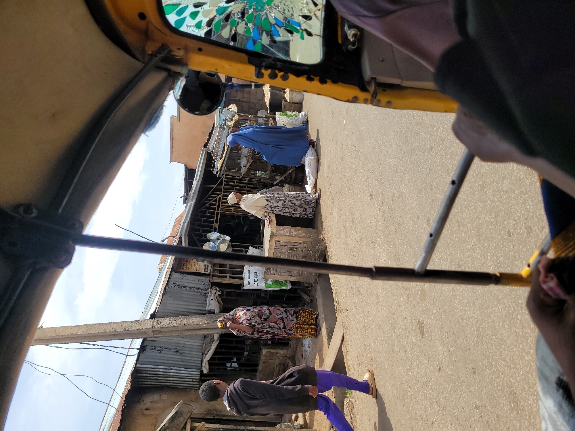 The image, taken from inside a yellow auto-rickshaw (keke) in Jos, Nigeria, shows a street scene. The street is paved and lined with informal shops under corrugated metal roofs. Several people are present: one in a long blue robe and head covering is bending over a large white sack; another, wearing a patterned dress, is standing near a shop; another is wearing a white head covering and a printed dress and is bending over what appears to be merchandise. One person in a dark gray jacket and purple pants is walking in the foreground. The auto-rickshaw’s frame and the driver's hands are visible in the foreground. The rickshaw's windshield has decorative elements.
