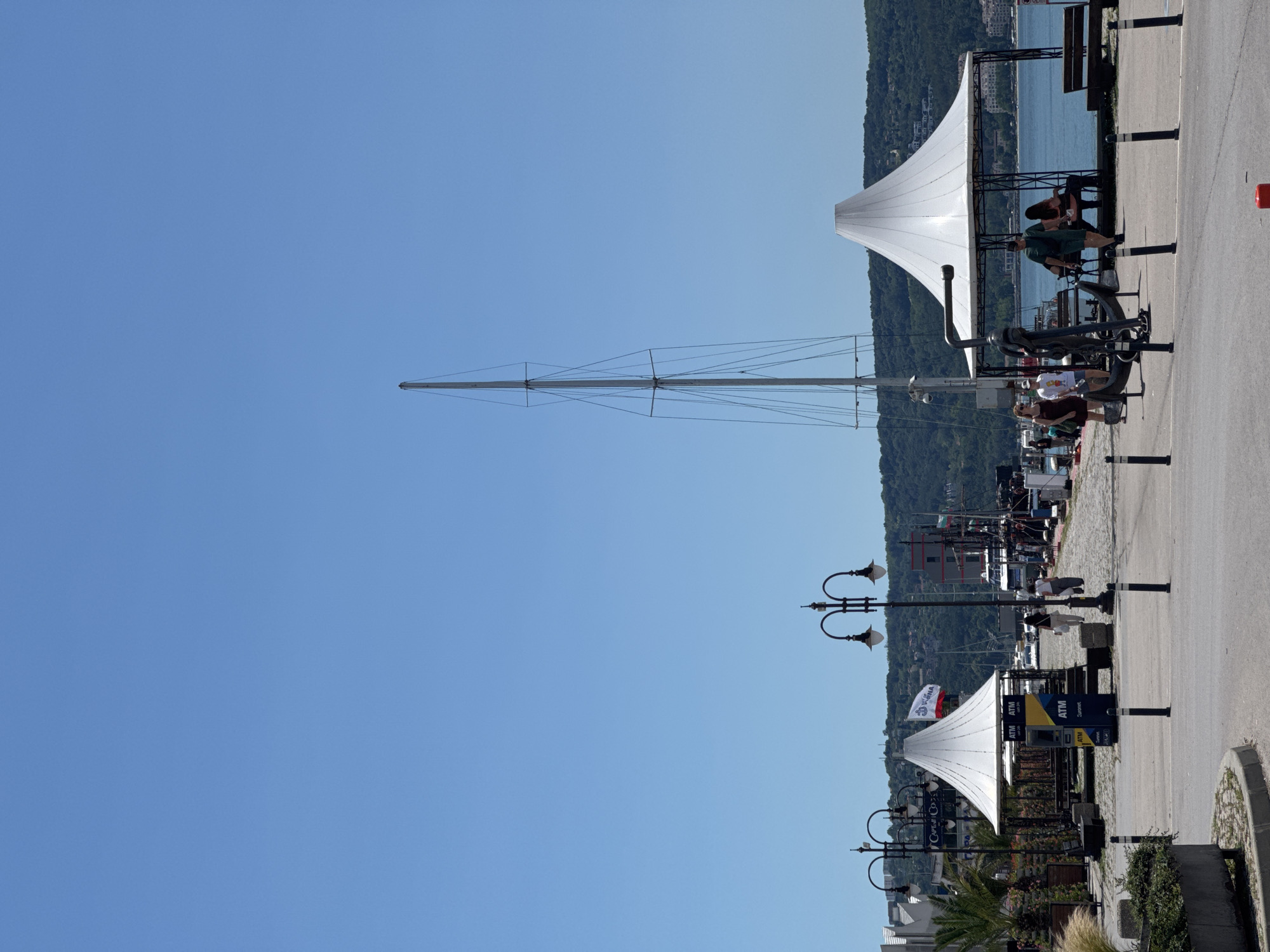 The image depicts a waterfront scene in a city with a clear blue sky overhead. In the foreground, a paved walkway runs parallel to the water, bordered by bollards. Several people are walking and sitting in this area. To the left, there's a white tent-like structure with seating underneath and planters with greenery. Further back, another, larger white tent structure is visible, with ATMs positioned in front of it. A tall, slender mast of a boat dominates the middle ground, with its rigging visible against the sky. Beyond the mast, a body of water is present, with several boats docked and moored. In the background, a lush green hillside covered with trees and buildings rises above the waterfront. The overall impression is that of a lively, sunny day at a harbor or marina.