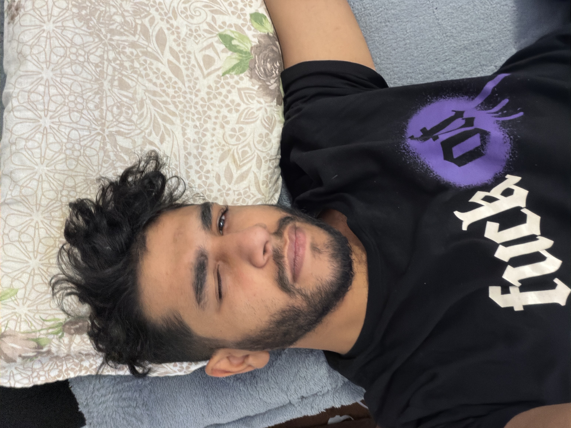 A medium shot shows a man with dark, curly hair and a beard winking at the camera. He is lying on his side, resting his head on a beige and white patterned pillow with a green floral design in the corner. He is wearing a black t-shirt with a large, stylized white word and a purple emblem on the upper left. The man appears to be on a gray blanket or cushion. The location is unknown, with no indication of being in Wirges, Germany.