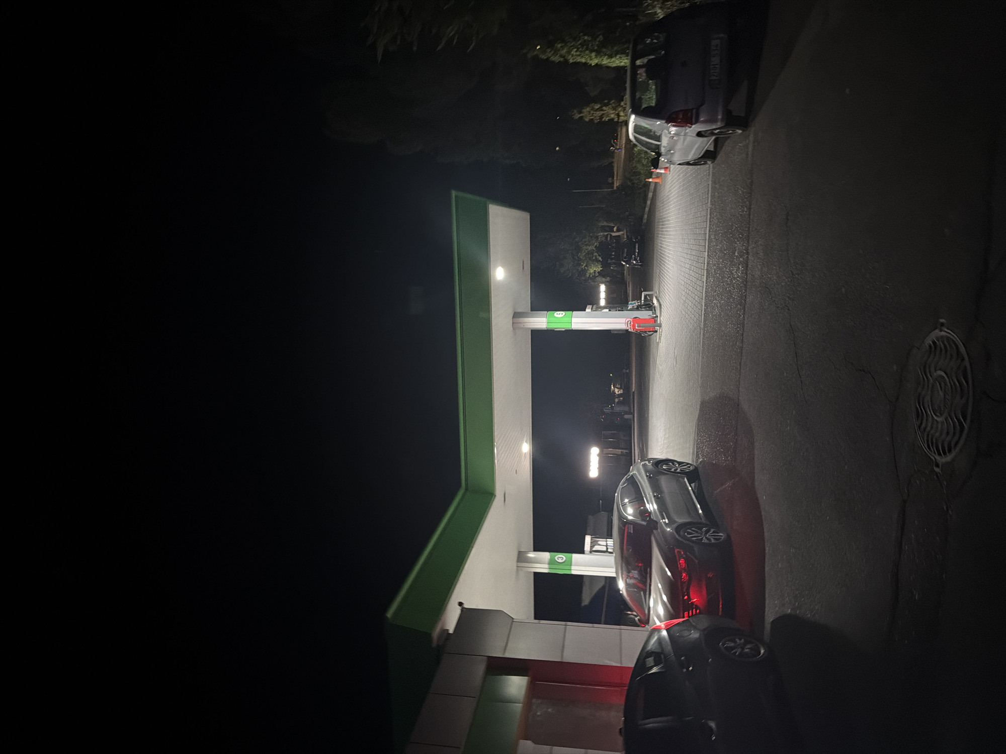 The image depicts a nighttime scene at a gas station, likely located in Chișinău, Moldova. Several cars are present: a dark-colored sedan is parked to the left, a silver sedan is positioned in the center, and a small silver hatchback is on the right. The gas station features a covered canopy with green trim, supported by white pillars. One pillar displays the number "3" in a green circle, and another shows a green "9". Red and blue gas pumps are visible. The ground is paved with asphalt and brickwork. Trees and foliage line the right side of the scene. The overall lighting is dim, with artificial lights from the gas station illuminating the area.