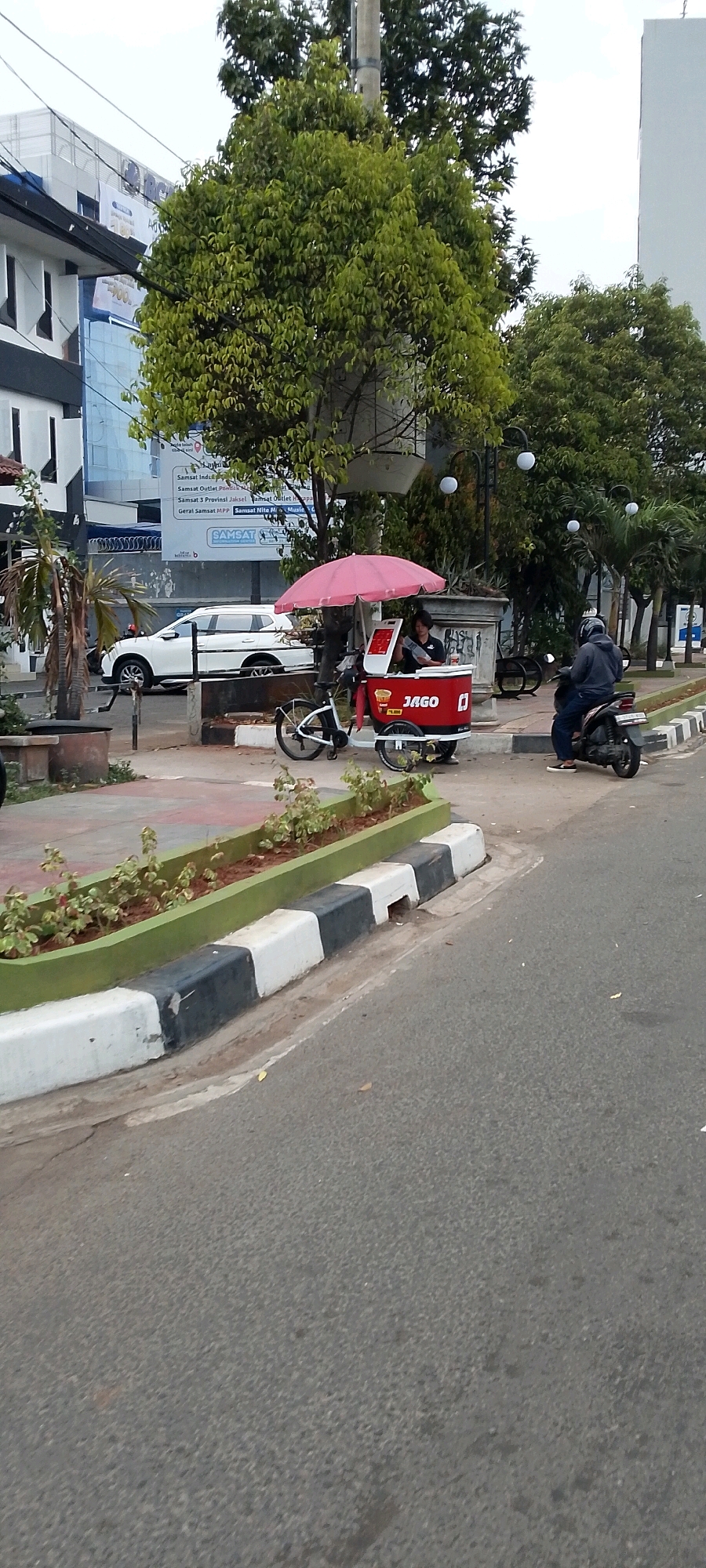 The image shows a street scene in Bekasi, Indonesia, with a white SUV parked on the left side. In the foreground, a vendor is operating a red and white cart, which is open at the top with a pink umbrella. The cart has the word "JAGO" printed on its side. A person is standing behind the cart, appearing to be serving a customer or preparing something. To the right of the cart, a person is riding a black scooter. The street has a paved surface with a curb that is painted with black and white stripes. A planter box with small green plants is situated along the curb. In the background, there are buildings and trees, with some power lines visible overhead. The sky is overcast.