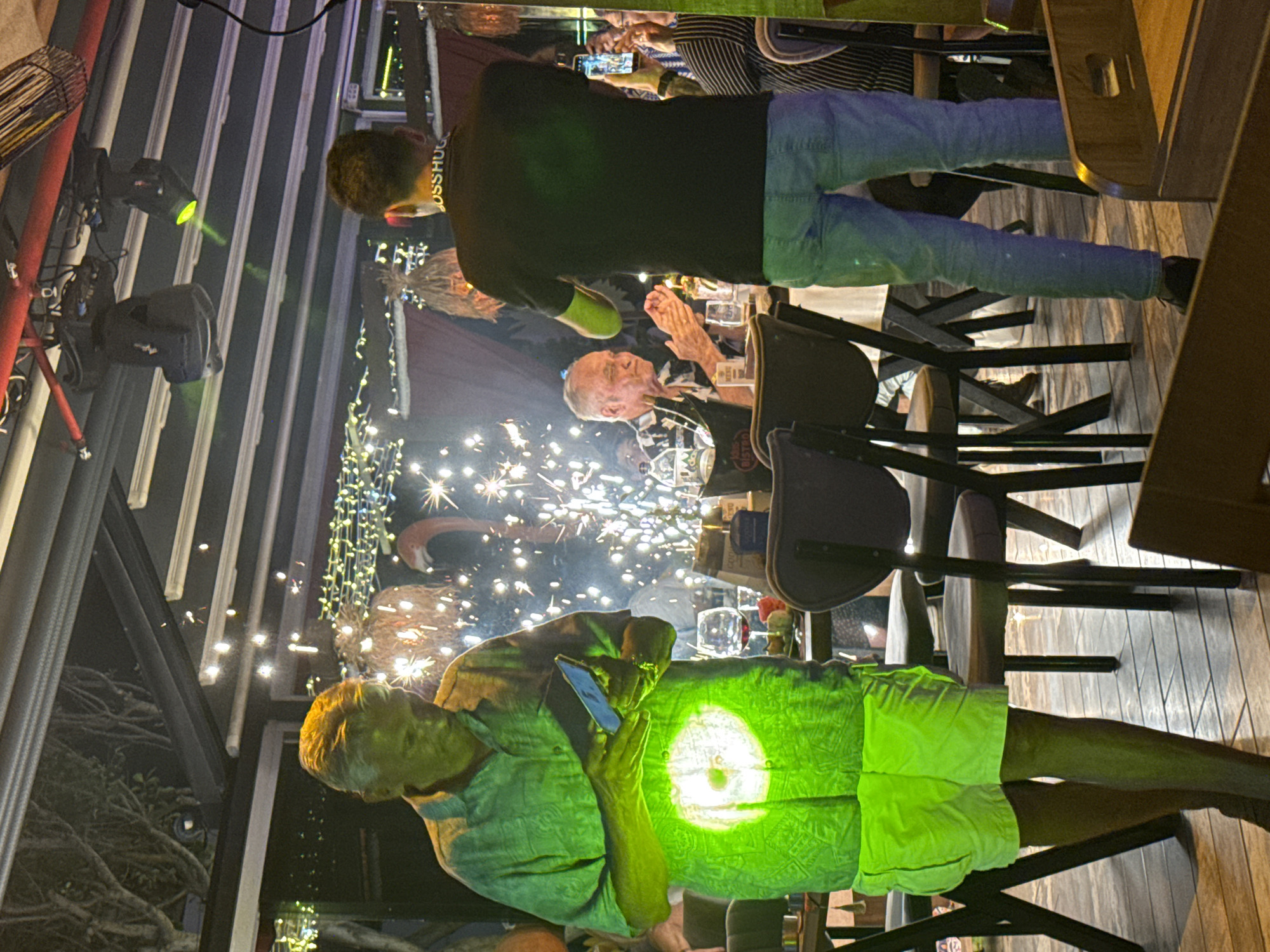 This image captures a nighttime scene inside what appears to be a lively restaurant, bar, or event venue with a covered ceiling. Several people are visible, illuminated by artificial light sources.
In the foreground, a man wearing a light green shirt and shorts holds a smartphone that emits a strong, vibrant green light, brightly illuminating his torso and lap. His legs are also visible, resting on what seems to be wooden flooring or decking.
Towards the center, another man, seen from behind, wears a black t-shirt, light blue jeans, and a necklace or collar with the word "BOSSHUG" visible on it. He is standing and looking towards the right side of the frame.
To the right, an older man with white hair is seated at a table, looking towards the left. His hands are raised as if he is clapping or gesturing. A partially obscured logo, possibly "BISTRO," is visible on his shirt.
The background features bright, erupting sparklers, suggesting a celebration or special occasion. Other patrons are partially visible further back, including one holding a smartphone, possibly capturing the event. The venue is adorned with decorative string lights and spotlights, some of which cast green beams. The overall ambiance is dim but festive, indicating a social gathering at night.