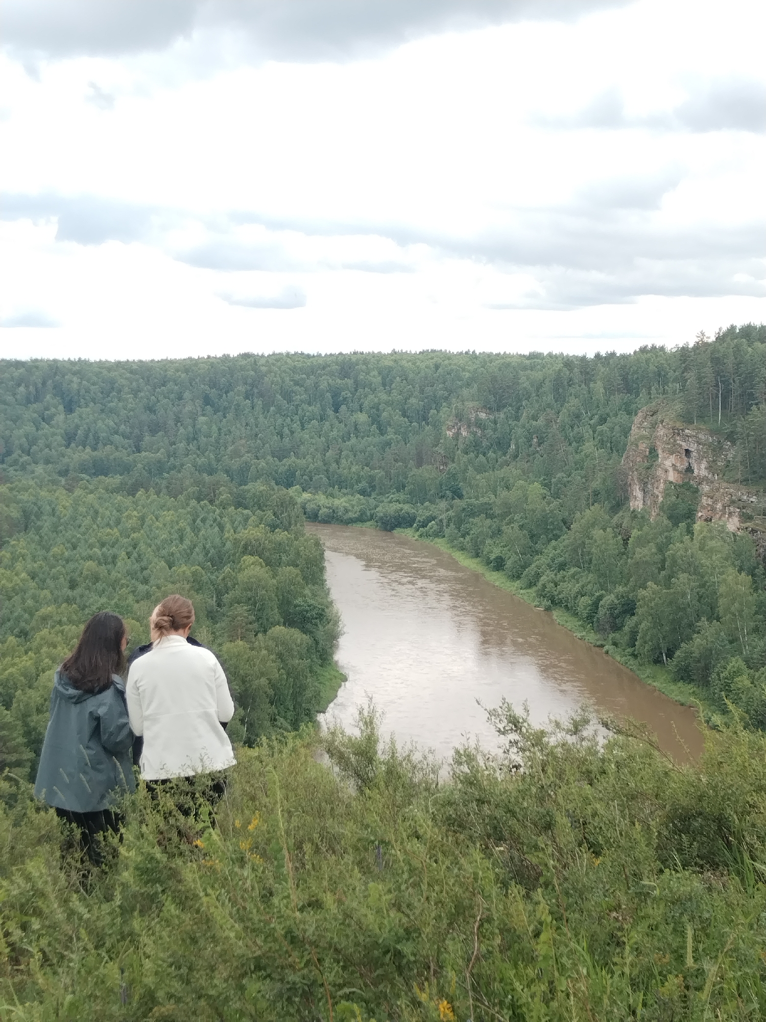 Two individuals, one wearing a dark teal rain jacket and the other a white jacket, stand on a grassy, overgrown hillside overlooking a wide, muddy river that winds through a dense forest. The forest covers rolling hills and cliffs, with a rocky outcrop visible on the right side of the frame. The sky is overcast with heavy, gray clouds. The scene appears to be a natural landscape, possibly a park or a viewpoint in the vicinity of Idrisovo, Russia, given the description.