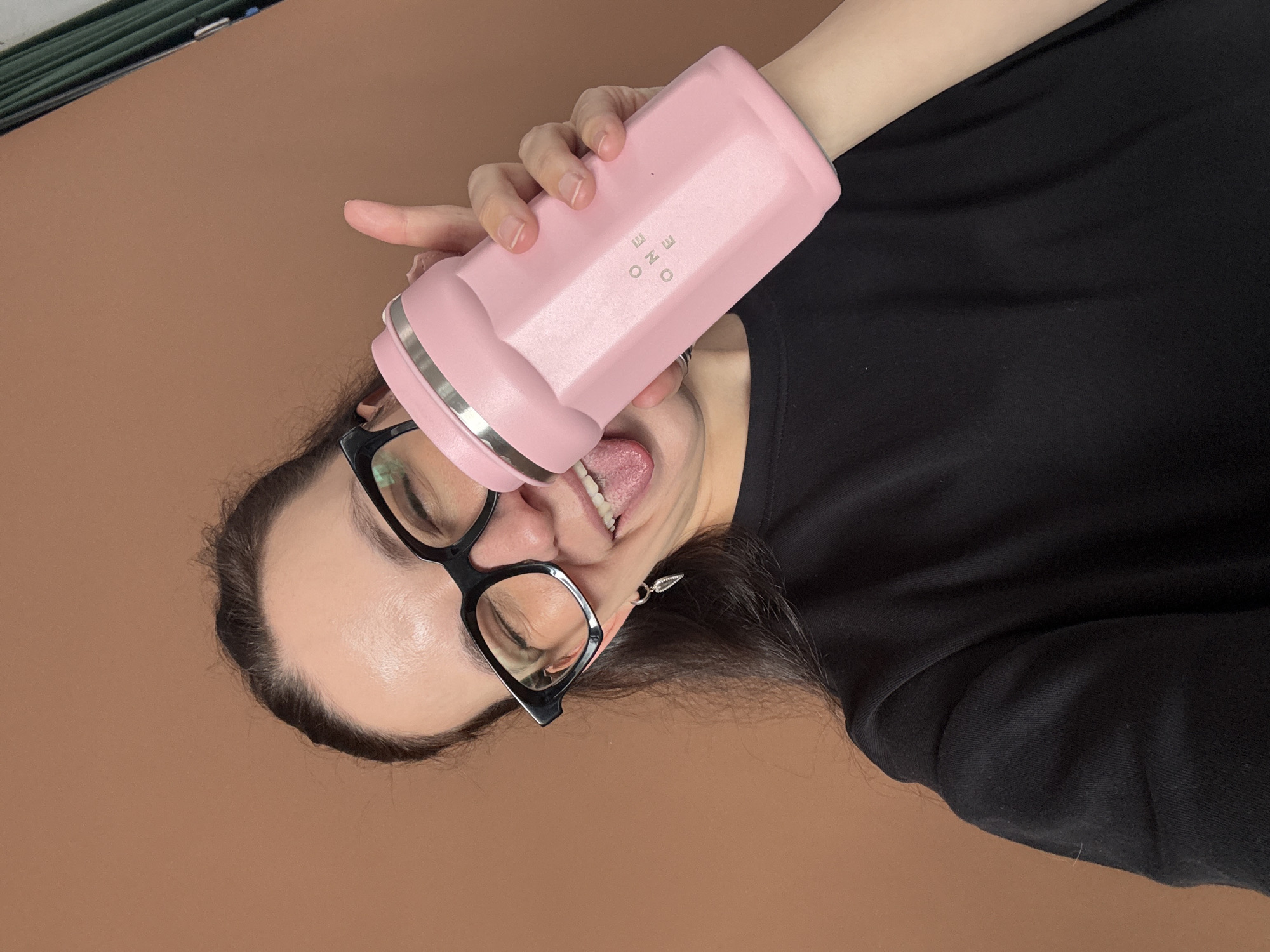 A young woman with dark hair and glasses is holding a pink water bottle to her mouth, sticking her tongue out playfully. She is wearing a black t-shirt and a silver earring. The background is a plain brown wall. There is no information available about the location being in Chaikovsky, Russia.