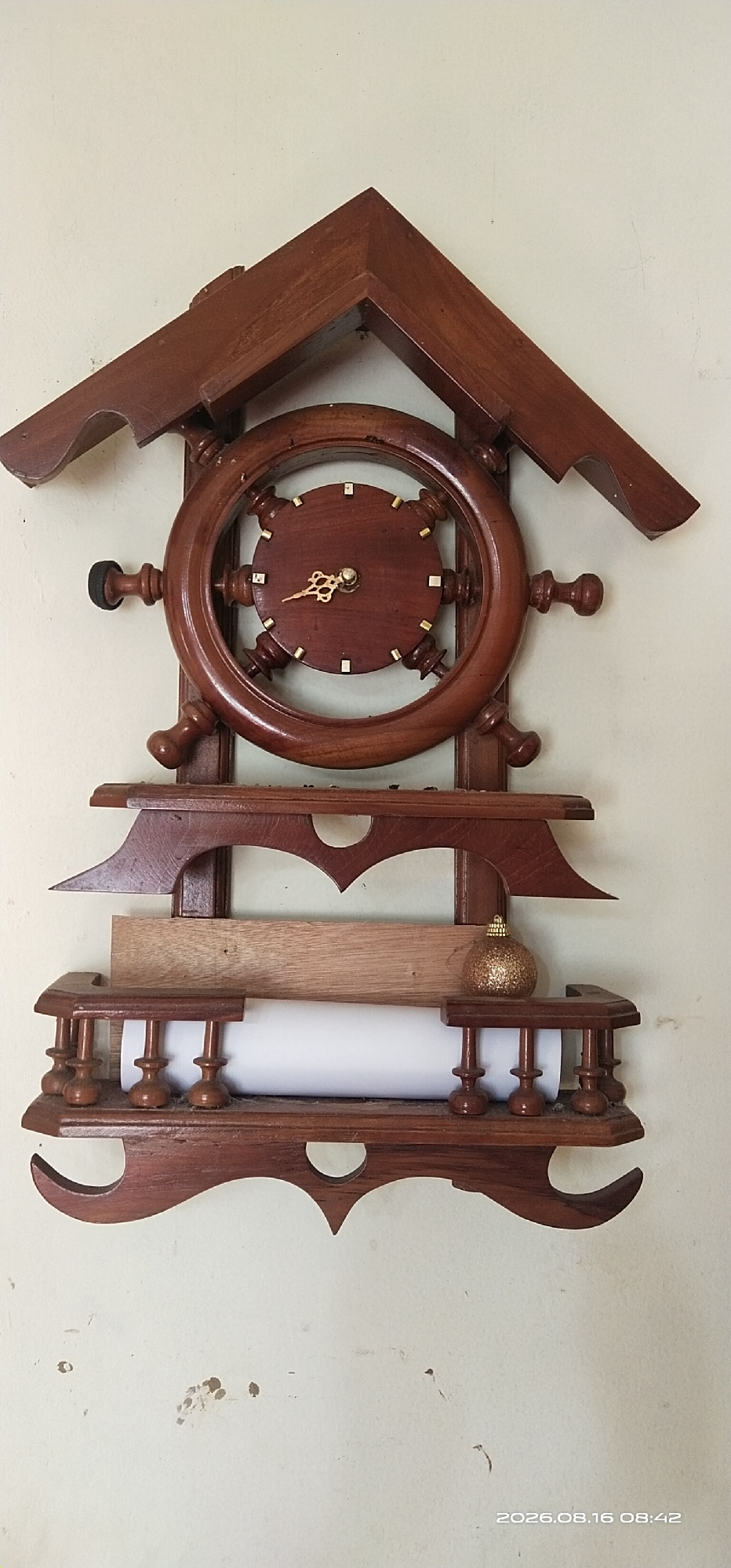 The image shows a wooden wall clock shaped like a ship's wheel, mounted on a pale, textured wall. The clock itself is made of dark wood with a circular face and gold-colored hands. It is surrounded by a larger wooden wheel structure with spokes. Below the clock, there are several wooden shelves and decorative elements. On one shelf, a roll of white paper is visible, with a small, sparkly gold ornament placed on top of it. A piece of light-colored wood is also present on a shelf above the paper roll. The wall shows some subtle staining and marks. The lower right corner of the image contains a timestamp: "2026.08.16 08:42". The scene appears to be indoors, likely a residential setting.