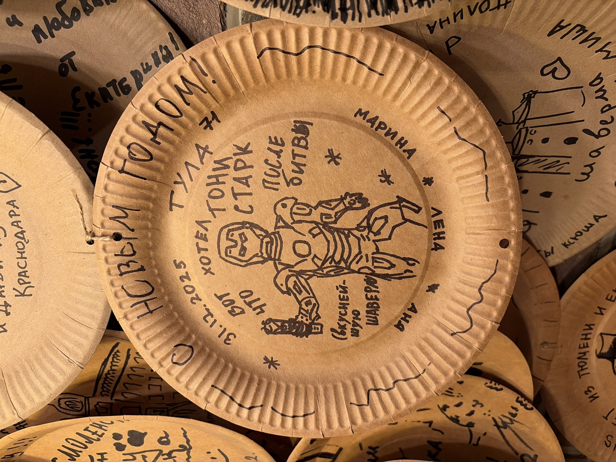 The image shows a collection of brown paper plates decorated with black marker drawings and handwriting. One plate in the foreground has a drawing of Iron Man and the text "С НОВЫМ ГОДОМ!!!" (Happy New Year!!!) written in a circle around it. Below Iron Man, there is text that appears to read "(вкуснейшую шаверму)" (the tastiest shawarma). Other text on this plate includes "31.12.2025", "ТУЛА", "ХОТЕЛ", "ТОНИ СТАРК", "ПОСЛЕ", "БИТВЫ", and "МАРИНА".
Other plates are visible in the background, also decorated with drawings and writing. One plate to the left has a drawing of buildings and text that includes "С МОЛЕНСКА". Another plate to the top has text including "КРАСНОДАРА". A plate to the right has text including "ЕКАТЕРИНБУРГ" and "ЛЮБОВЬ". The plates are arranged overlapping each other, suggesting they are part of a display or collection. The lighting is warm and appears to be artificial, creating shadows and highlighting the textures of the paper plates. The background appears to be a brick wall.