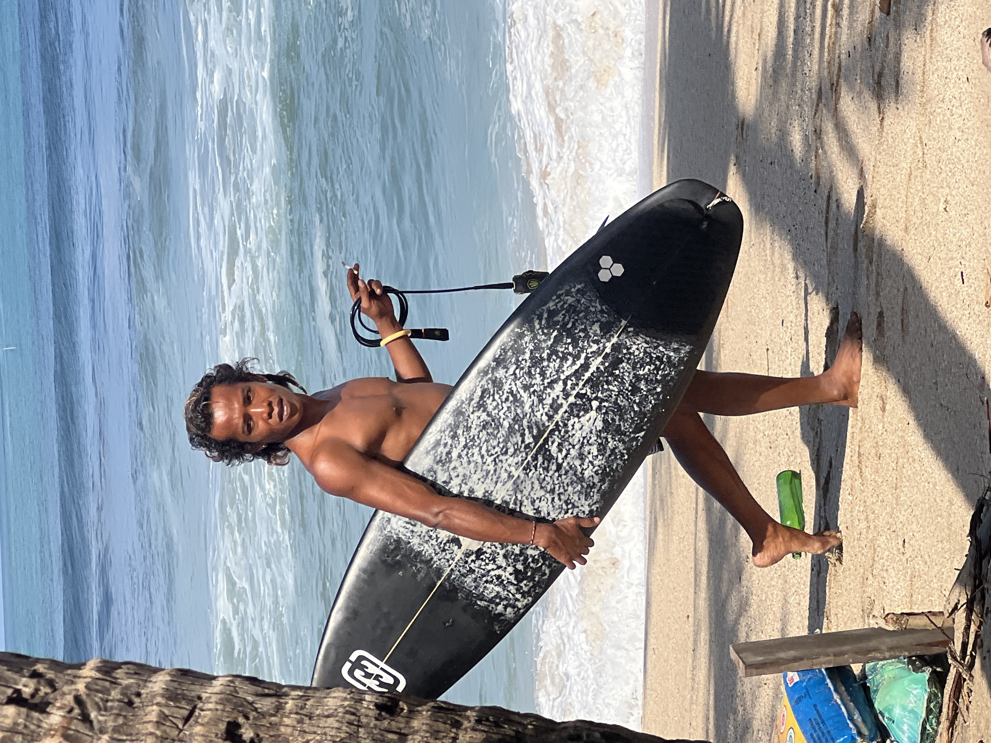 A shirtless man with dark, wavy hair walks along a sandy beach, carrying a black surfboard. The surfboard has a white, speckled design and a white logo near the nose. He is holding a cigarette in his right hand and a leash for the surfboard in his left. The ocean with white-capped waves is visible behind him. The background shows the blue ocean and a clear sky. The scene is set in Kuta, Indonesia.
