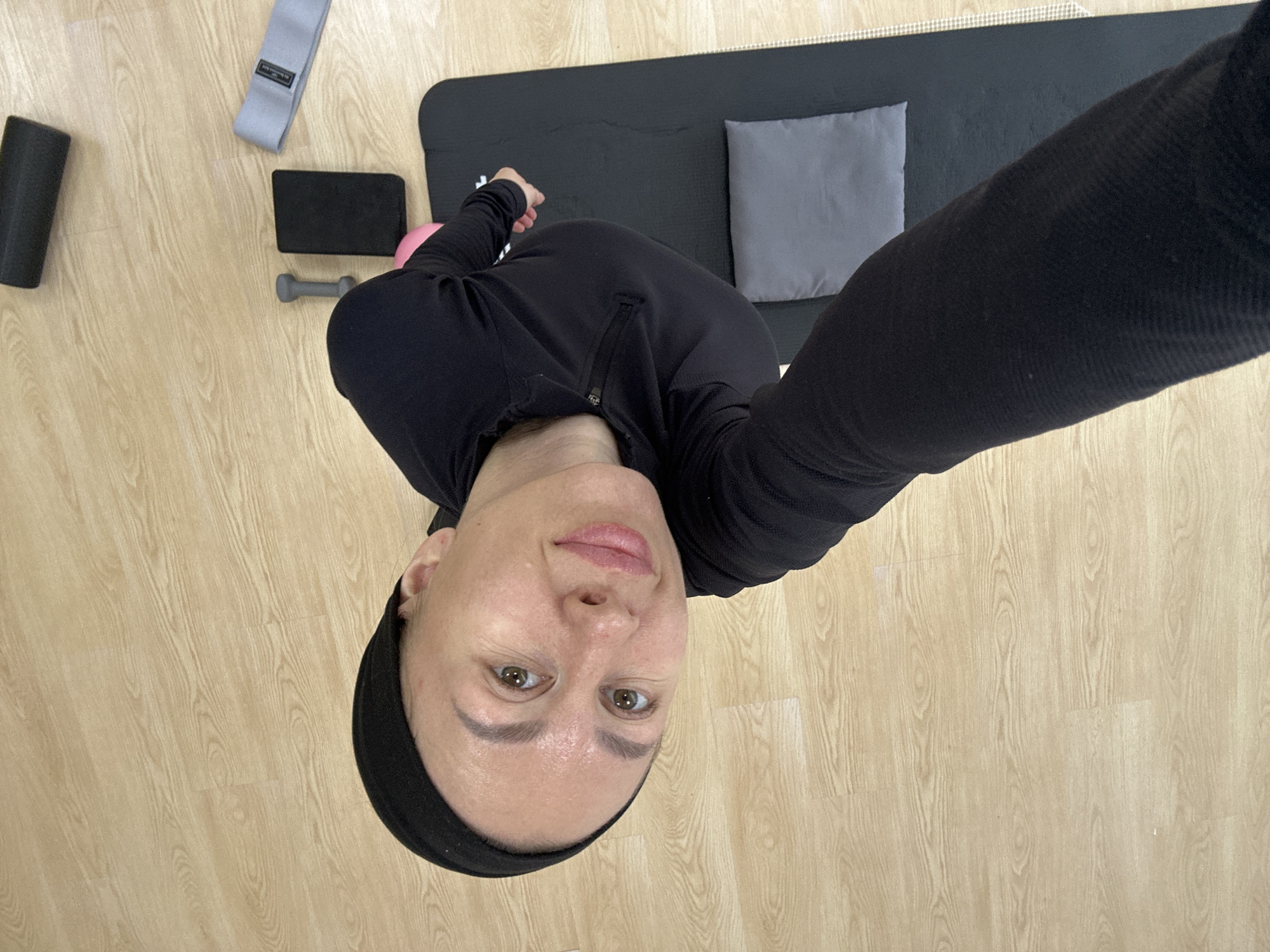 A person wearing a black headband and black long-sleeved top is angled towards the camera in an indoor setting, likely a gym or home exercise space. A black exercise mat lies on the floor, along with a gray pillow, a black foam roller, a gray resistance band, a black block, a small pink ball, and a gray dumbbell. The floor is light wood-toned. The location is not discernible from the visual information provided.