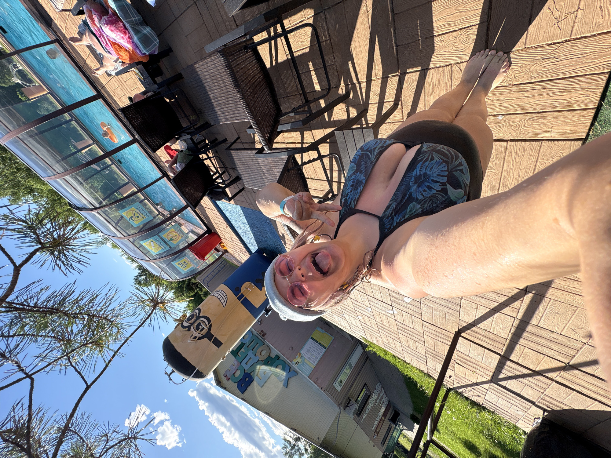 The image depicts a sunny outdoor scene, likely at a recreational facility. In the foreground, a person in a dark and blue tropical-print swimsuit, a light blue beanie, and pink sunglasses poses for a selfie, sticking out their tongue. Their arms and torso are visible, with water droplets indicating they have recently been in the water.
Behind the person is a wooden deck, some of which is in shadow. To the left, a building with "BOGEET XOPOMU" (likely a Russian phrase) written on it is visible, along with a tall, cartoonish figure resembling a gas tank.
Further back, a covered swimming pool with blue water is present. Inside the pool, one person is visible on an orange inner tube, and another person is lying on a lounge chair near the pool, covered with colorful blankets. Several lounge chairs and small tables are placed on the deck around the pool. Trees and a bright blue sky with scattered clouds form the background. The overall environment suggests a warm, leisure-oriented setting.