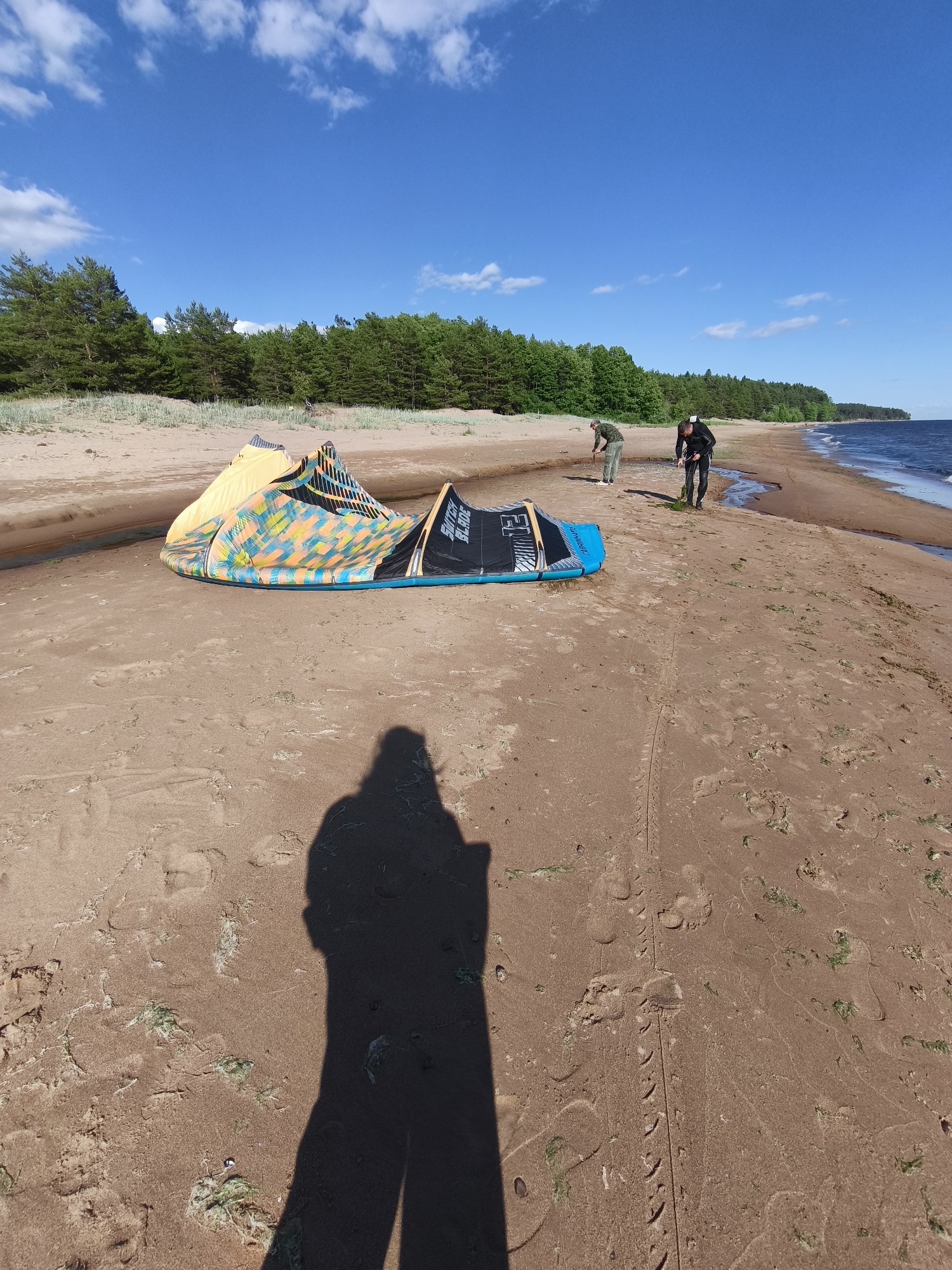 This media file depicts a sunny day at Zelenaya Roshcha, Russia, with a clear blue sky and scattered white clouds. The scene is a sandy beach bordering a calm body of water, with a dense forest of pine trees lining the horizon. In the foreground, two kitesurfing kites are laid out on the sand. The kites are colorful, one primarily yellow with black and blue accents, and the other is blue and black with white lettering.
Two individuals are present on the beach. One, wearing a dark outfit, appears to be preparing or has just finished a water activity, standing near the water's edge. The other person, dressed in lighter clothing, is further up the beach. A prominent shadow of the photographer, facing away from the viewer and towards the kites, is cast on the sand.
The presence of kitesurfing equipment suggests an activity related to kitesurfing or kiteboarding is either underway or about to begin. The overall atmosphere seems relaxed and geared towards outdoor recreation, typical of a pleasant day at the beach. The time of day appears to be late morning or early afternoon, given the bright sunlight and the long shadows. The sand is marked with footprints and some scattered green debris, possibly seaweed. Visible text on one of the kites reads "SWITCH BLADE".