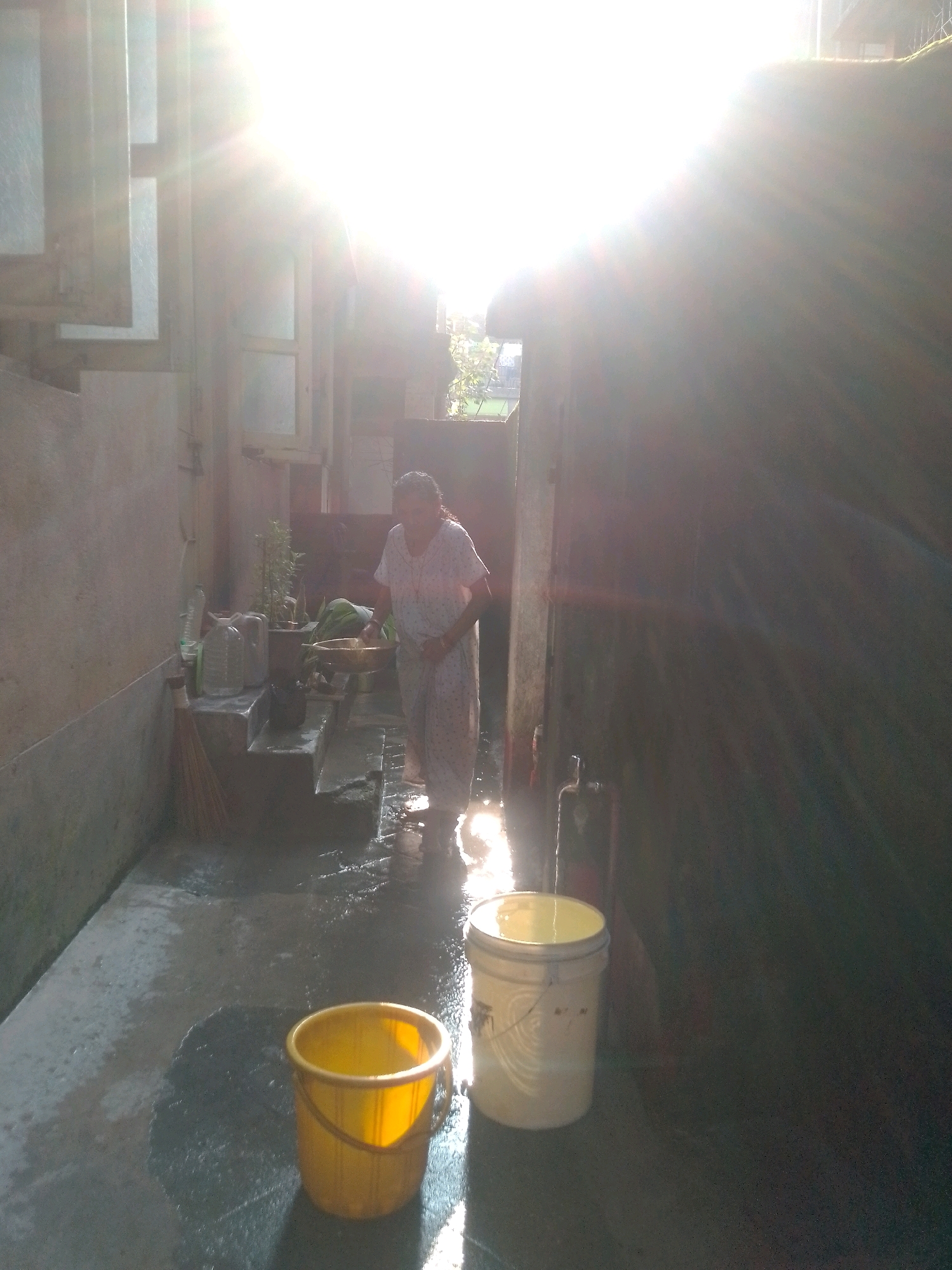 In Siliguri, India, a woman wearing a light-colored, patterned garment is standing on a wet, concrete surface in what appears to be an outdoor courtyard or wash area. She is holding a shallow, circular basin in her hands. Sunlight streams intensely from the upper left of the frame, creating lens flares and bright highlights. To her right, there is a large, pale-colored bucket. In the foreground, a bright yellow bucket is visible on the wet ground. Several water containers, including two large plastic jugs, are positioned on raised steps to the left of the woman, along with a broom leaning against the wall. The environment is lit by harsh sunlight, casting long shadows and creating a washed-out effect in some areas.