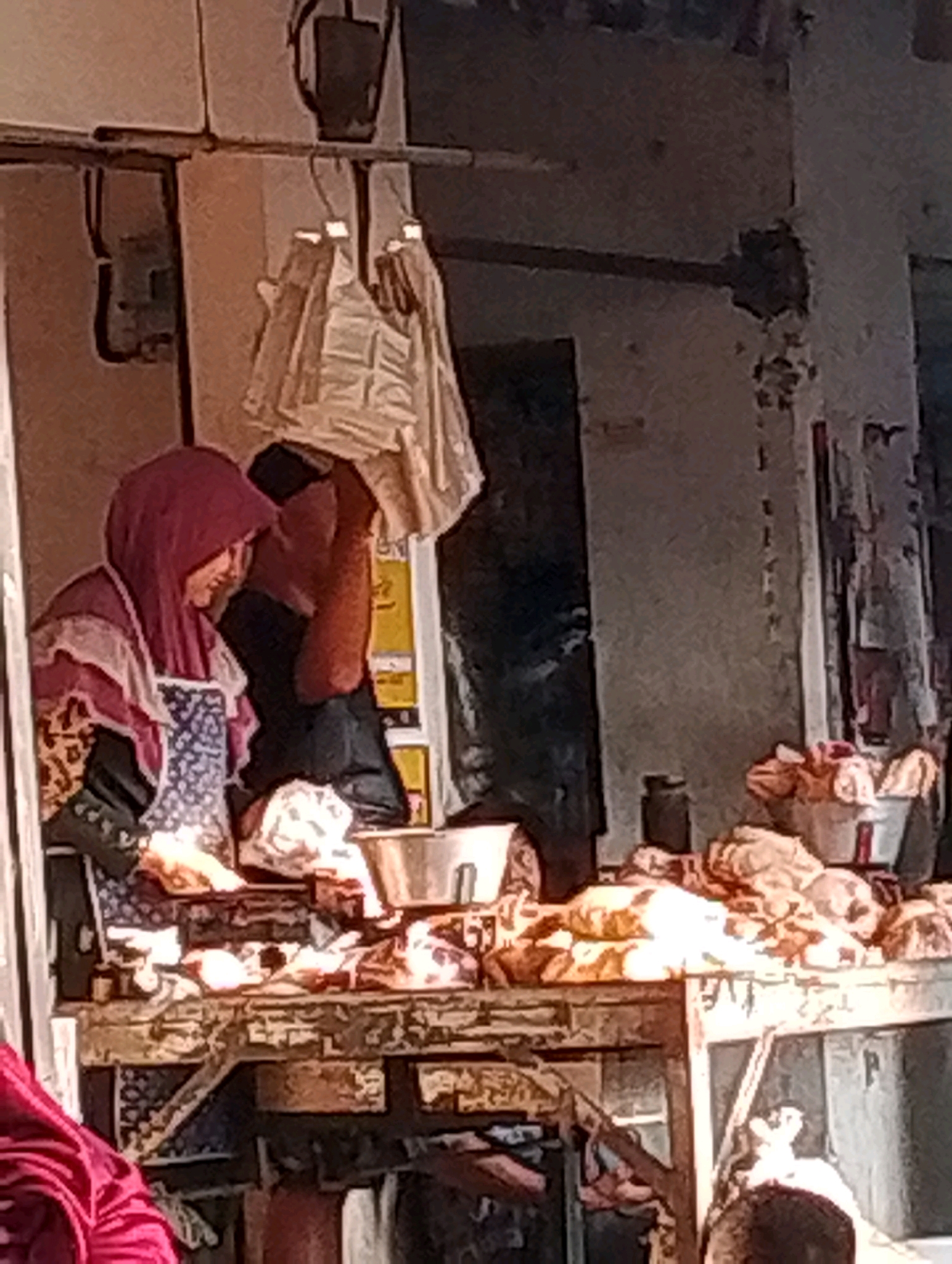 A woman wearing a pink hijab and an apron stands behind a wooden counter filled with meat. She is looking down at the meat, possibly preparing it. There are other people in the background, obscured by shadows and the general blurriness of the image. Bags of goods hang from a rack above. The scene appears to be a market stall. The image is of low quality and is pixelated, making it difficult to discern finer details. The location is identified as Probolinggo, Indonesia.