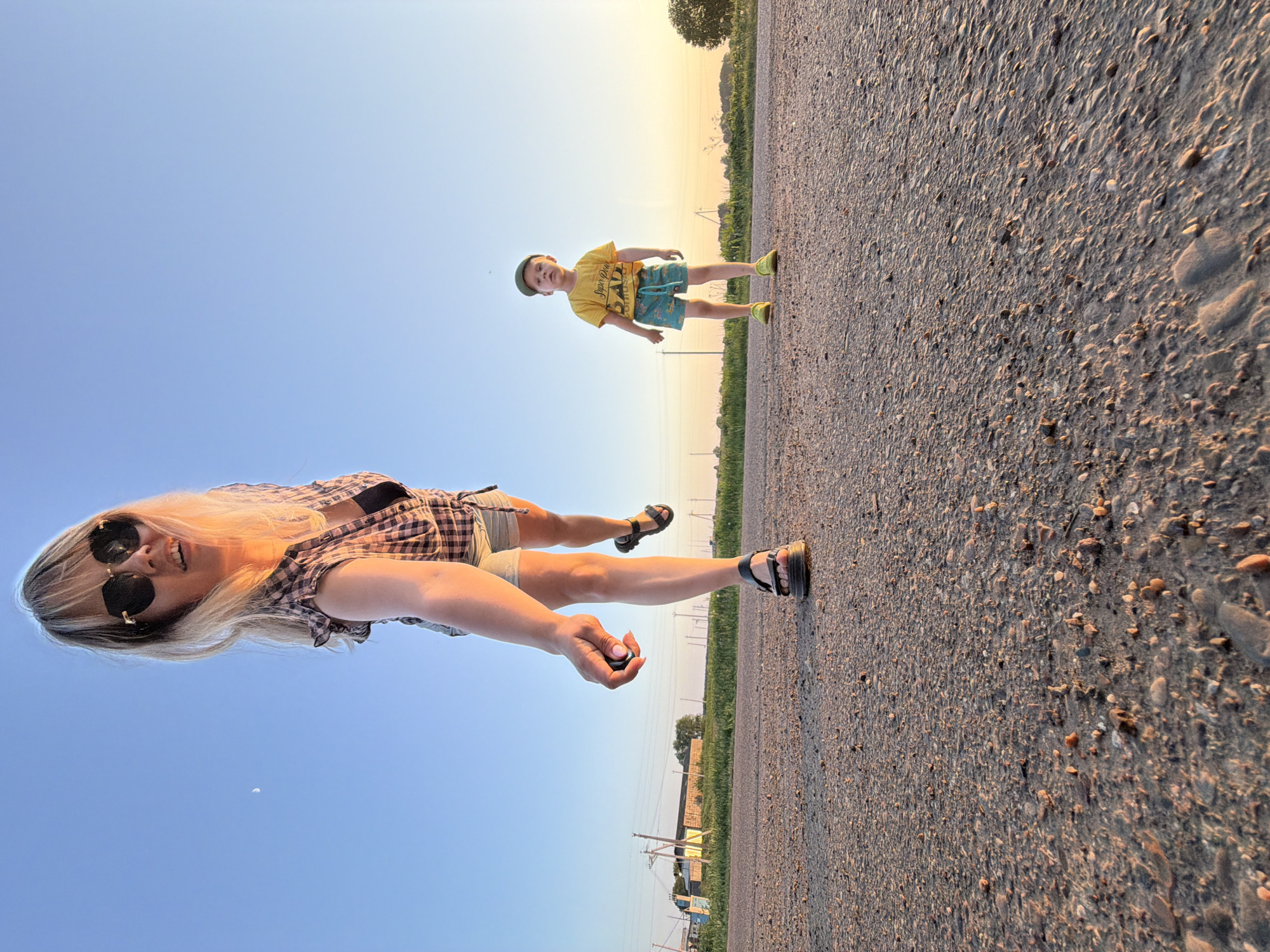 The image, taken in Чумна, Russia, presents an unusual perspective. A woman with blonde hair and sunglasses stands on a paved surface, possibly a road, facing the camera. She wears a plaid shirt and shorts. In the distance, a young boy in a yellow shirt and patterned shorts also stands on the road. The background transitions from the paved surface to grass and low-lying buildings. The sky is blue with a hint of sunset glow. The photo's perspective makes it appear as if the woman and boy are standing on the sky.
