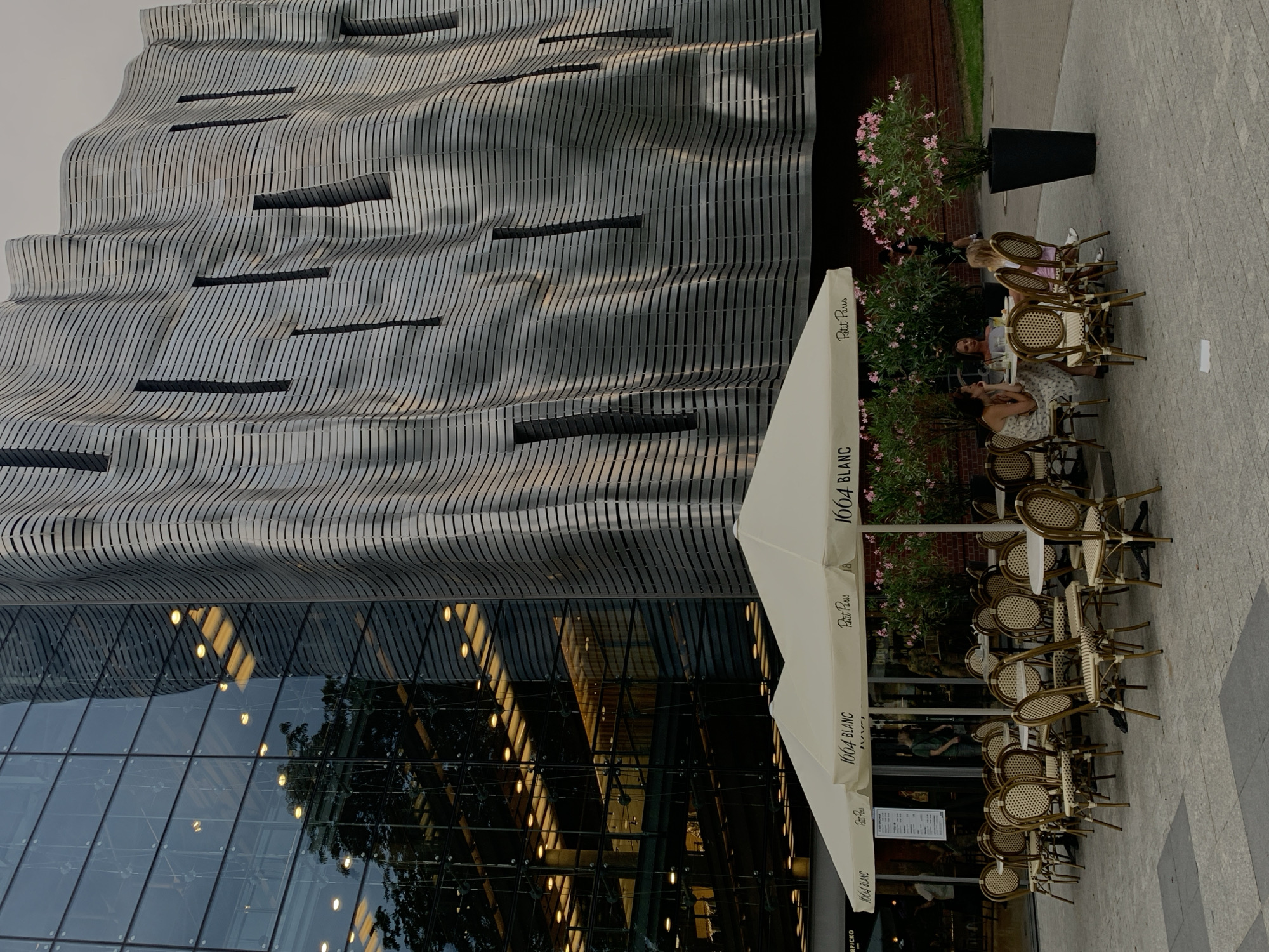 The image depicts an outdoor seating area of a restaurant or cafe in Poznan, Poland, situated in front of a modern building with a glass facade and a distinctive wavy, metallic exterior. Several white umbrellas, branded with "1604 BLANC" and "Petit Paris," provide shade over tables and chairs. The chairs are made of wicker and appear to be arranged around circular and square tables. Some of the tables are occupied by patrons. On the right side of the image, a large potted plant with pink flowers stands near the building. The ground is paved with square tiles. The building's glass facade reflects the sky and interior lights. The metallic exterior of the building features horizontal slats and vertical openings.