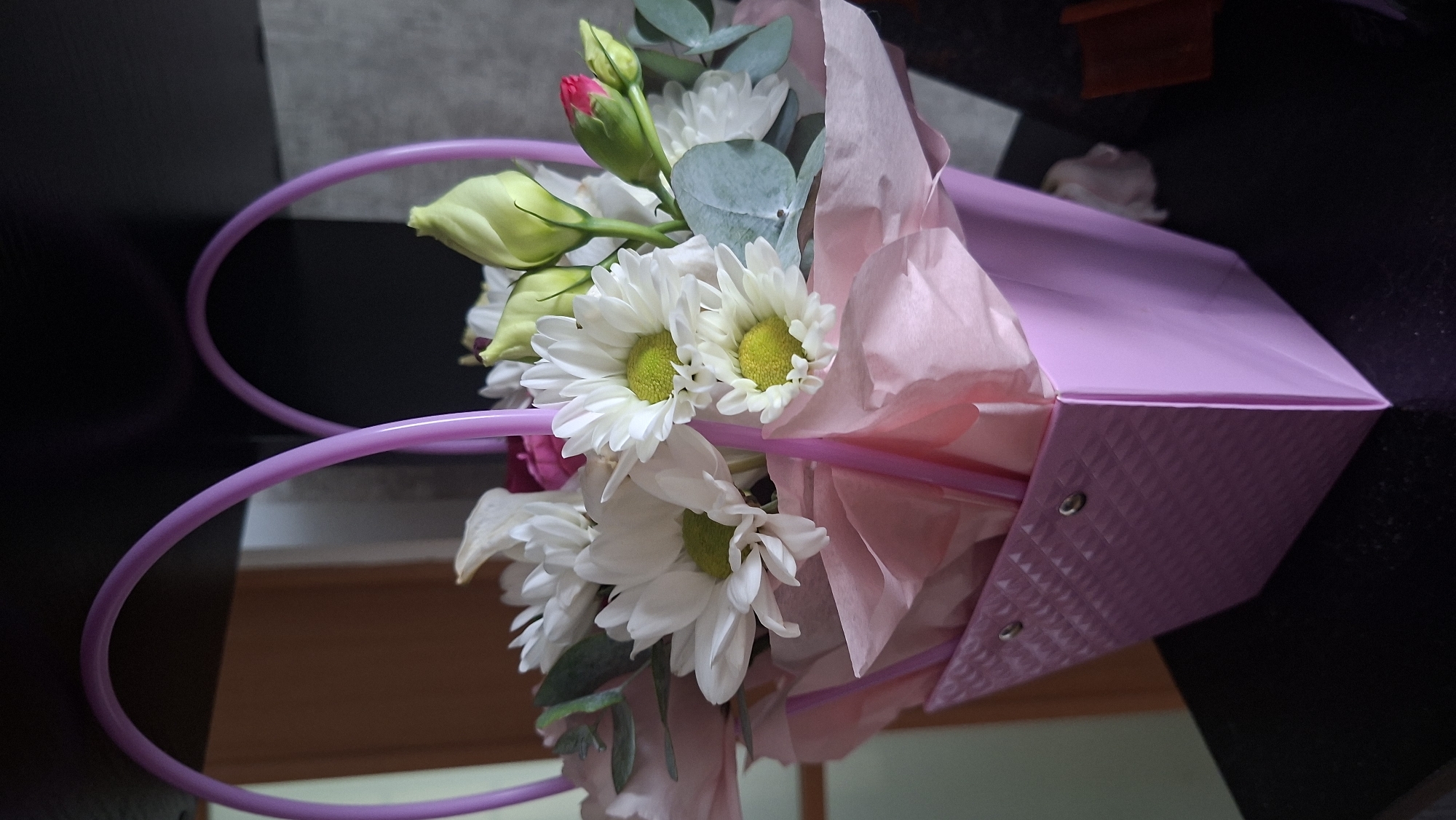A close-up, eye-level shot features a bouquet of white chrysanthemums and green buds in a pink textured gift bag with purple heart-shaped handles. The bag is adorned with silver grommets. The flowers and bag rest on a dark surface, possibly a table, with a blurred background that includes a light brown door and a gray wall. The scene appears to be indoors, in a domestic setting. There are no people or animals visible in the image. The lighting suggests an indoor environment with ambient light. No specific location in Myadzyel, Belarus can be determined from the visual information provided.