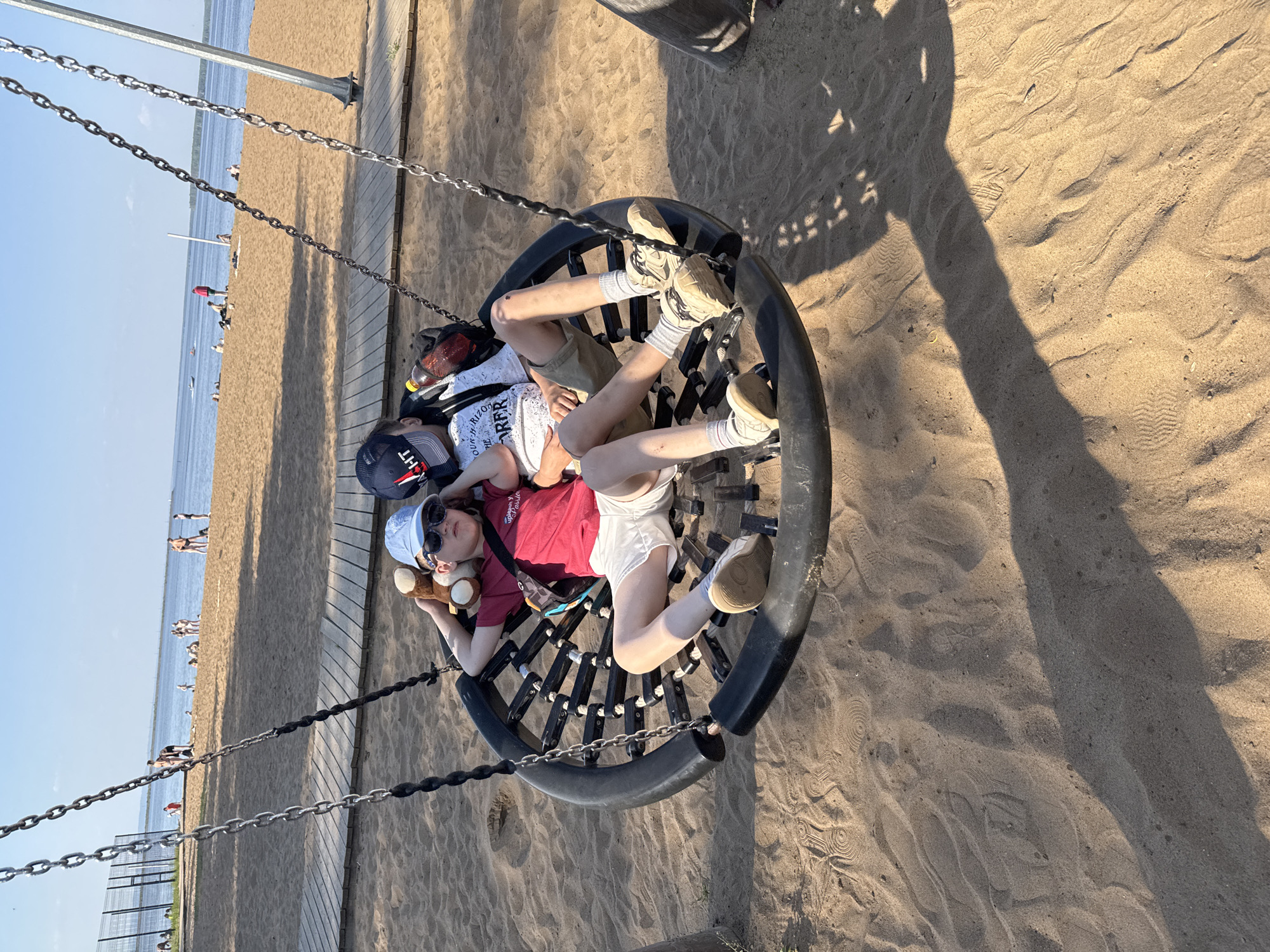Two children are lying in a large, round swing on a sandy beach. One child, wearing a red shirt, white shorts, and a blue baseball cap, is holding a stuffed animal. The other child is wearing a white shirt and khaki shorts. The swing is suspended by chains from a metal frame. The beach is populated with people in the background. The scene is located in Chaikovsky, Russia, as inferred from the location of the photographer.
