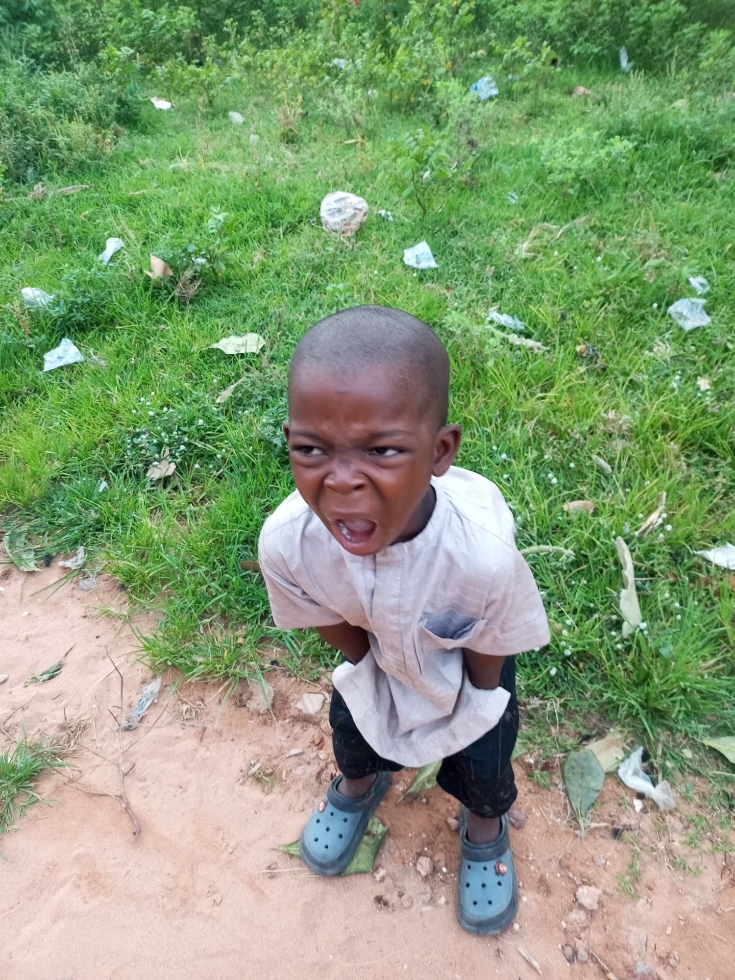 A young boy stands in a sandy area with sparse green grass and some scattered debris. He is wearing a light-colored tunic and dark pants, and blue clogs on his feet. The boy is looking directly at the camera with his mouth open in a grimace or shout, his eyes wide. The background consists of dense green foliage and more scattered debris, typical of an outdoor environment in Tempure, Nigeria. The lighting suggests it is daytime.