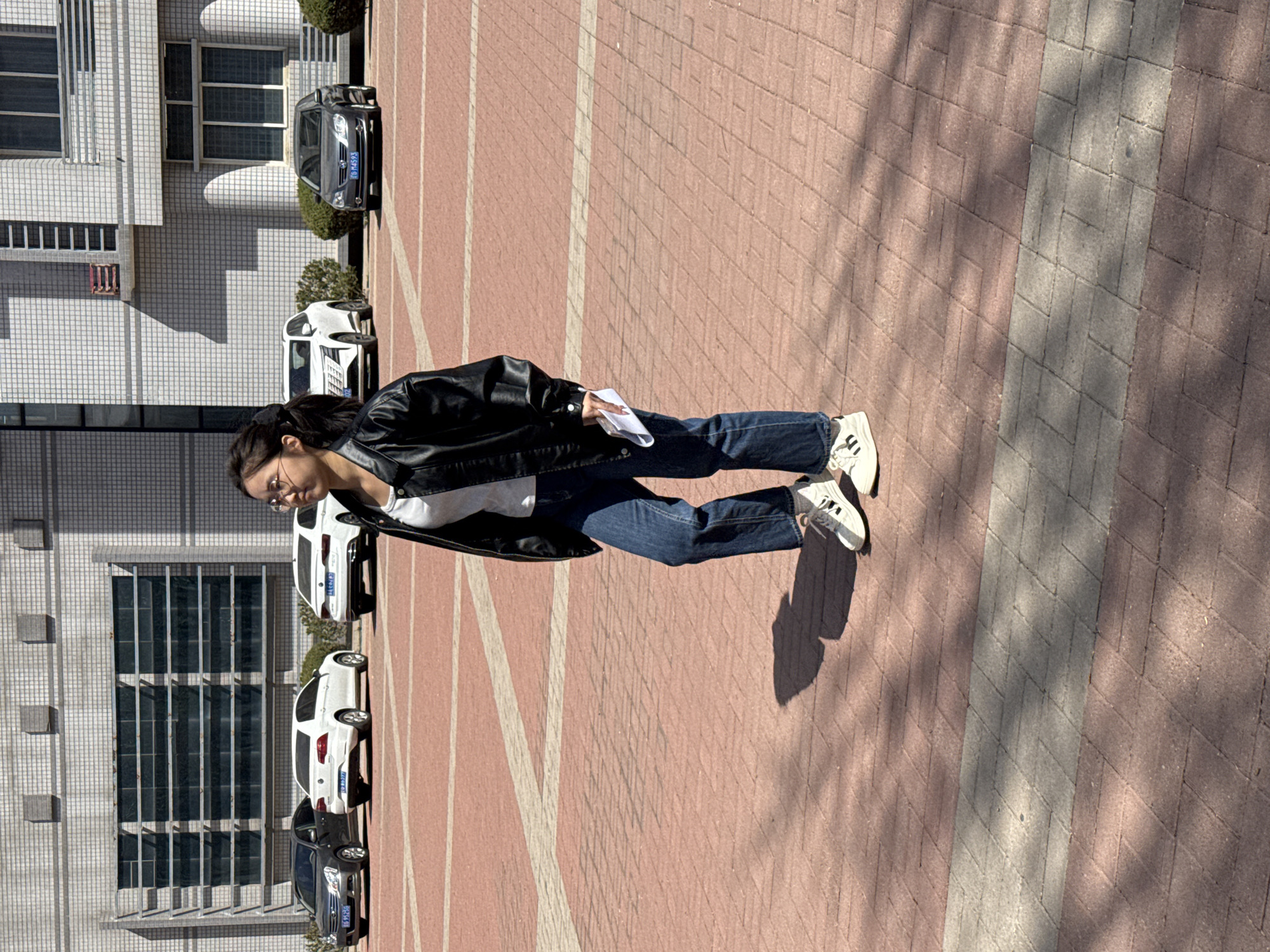 The image depicts a young woman standing outdoors on a bright, sunny day in Zhongtun, China. She is wearing a black leather jacket, a white t-shirt, blue jeans, and white sneakers with black stripes, and glasses. She holds a piece of paper in her hands, looking downwards.
The setting is an open paved area with reddish-brown and light grey rectangular interlocking bricks forming patterns on the ground. Distinct shadows from the woman, unseen trees, and buildings are cast across the pavement, indicating strong daylight, likely mid-morning to afternoon.
In the background, a multi-story building with a tiled facade and numerous large windows runs along the side. Several cars are parked parallel to the building. Visible vehicles include a dark grey sedan, a white SUV, another white sedan, and a black sedan. Car license plates with Chinese characters are visible; one plate clearly reads "鲁D M4593", and others show "鲁D", identifying them as vehicles from Shandong province, China. Small shrubs are planted near the parked cars. The scene appears to be an urban environment or a university campus.