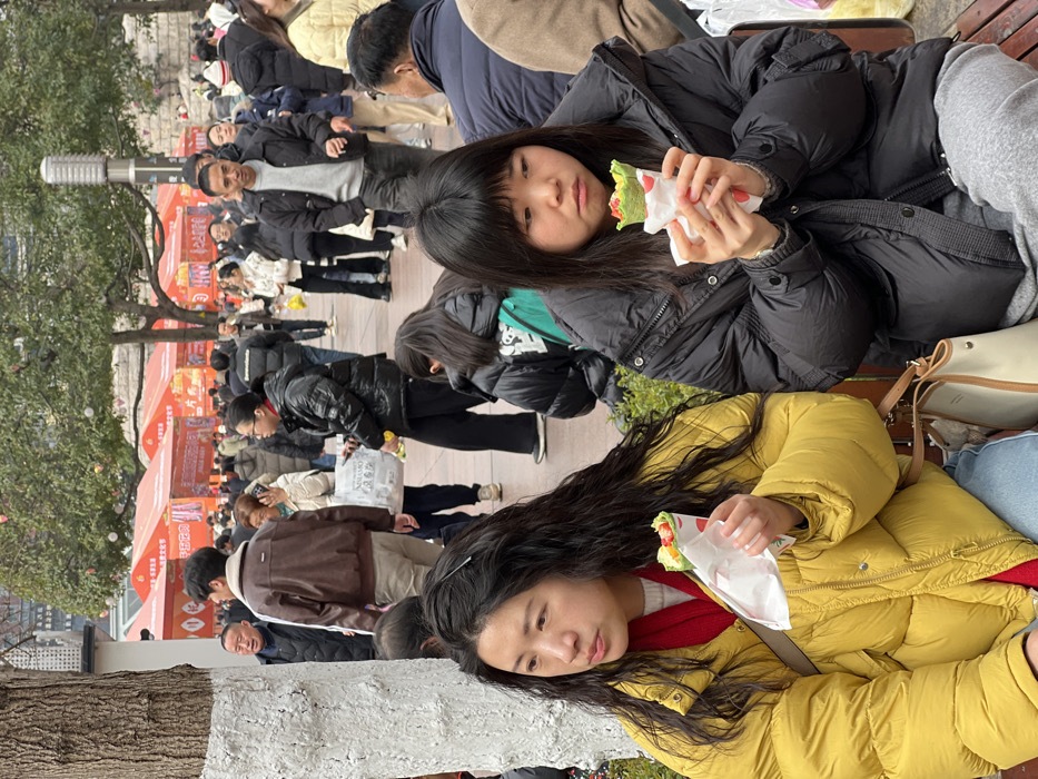 Two young women sit close together on a bench, each holding a wrapped sandwich. The woman in the foreground wears a yellow puffy jacket and has long, dark, wavy hair. The woman behind her wears a black puffy jacket and has short, straight, dark hair with bangs. A crowd of people mills about in the background, with vendor tents visible. The bench appears to be in a park-like setting with trees and a light pole nearby.
