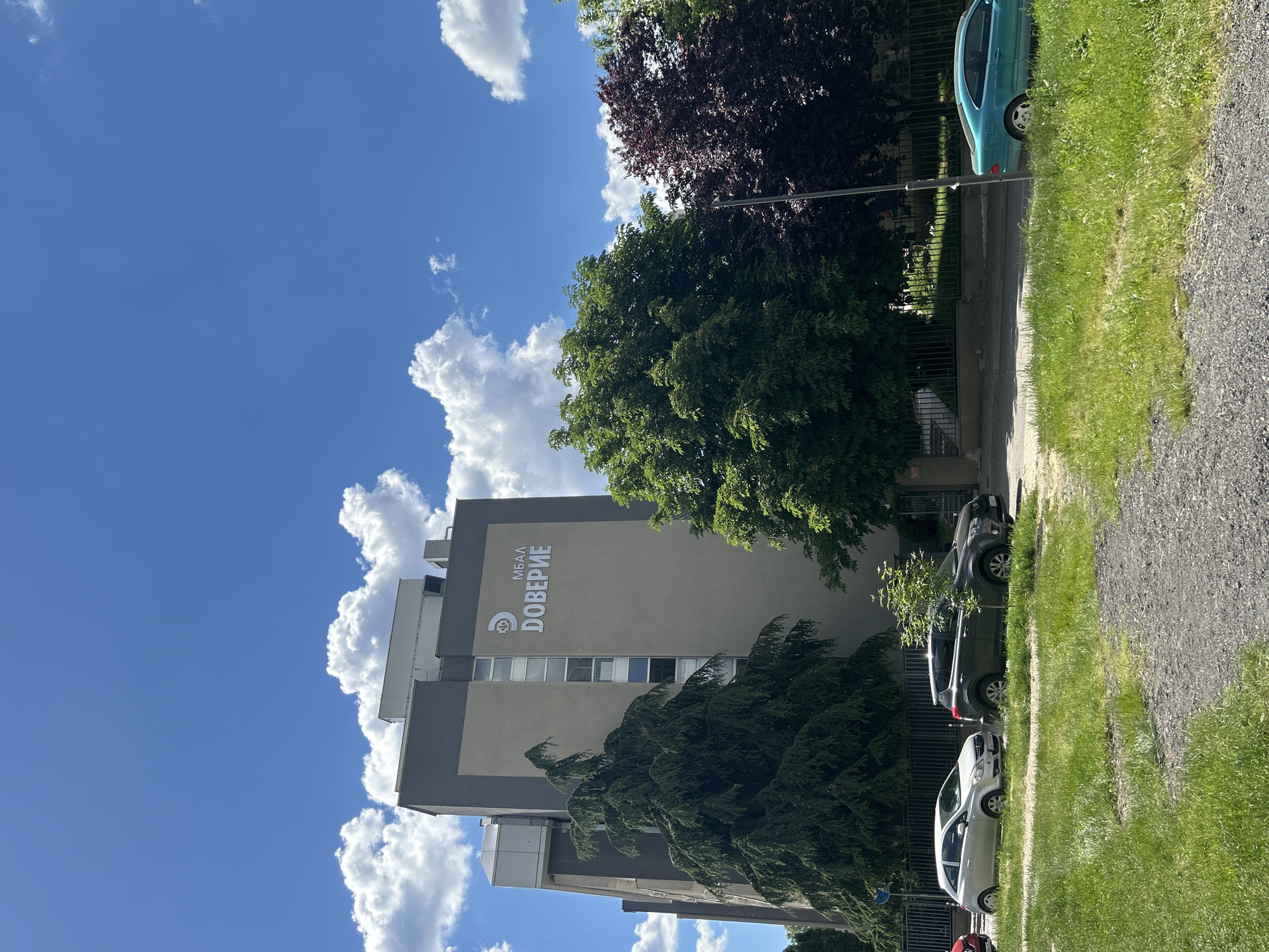The image shows a building with the text "МБАЛ ДОВЕРИЕ" on its side, located in an urban area. A blue sky with white clouds is visible in the background. Trees and parked cars are present along the side of the building. A grassy area and a gravel path are in the foreground. The city and specific location are unknown.