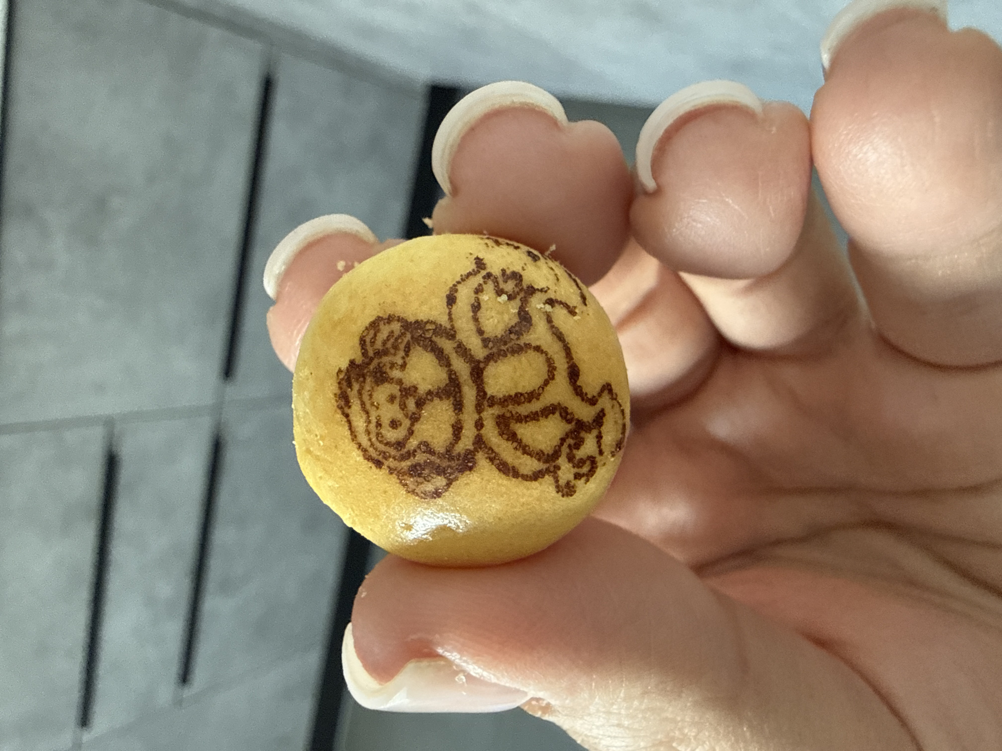A close-up shot shows a person's hand holding a small, round, yellow cookie or pastry with a brown drawing of a monkey on it. The monkey drawing is simple and appears to be done with edible ink or by pressing into the dough. The cookie has a slightly textured surface. Several fingers, with manicured nails, are visible around the cookie, supporting it. The background is out of focus but appears to be an indoor setting with a textured gray wall with horizontal lines, possibly cabinetry or a wall finish. The lighting is bright, highlighting the texture and color of the cookie and skin. There is no information in the image that can place this scene in Lyubertsy, Russia.