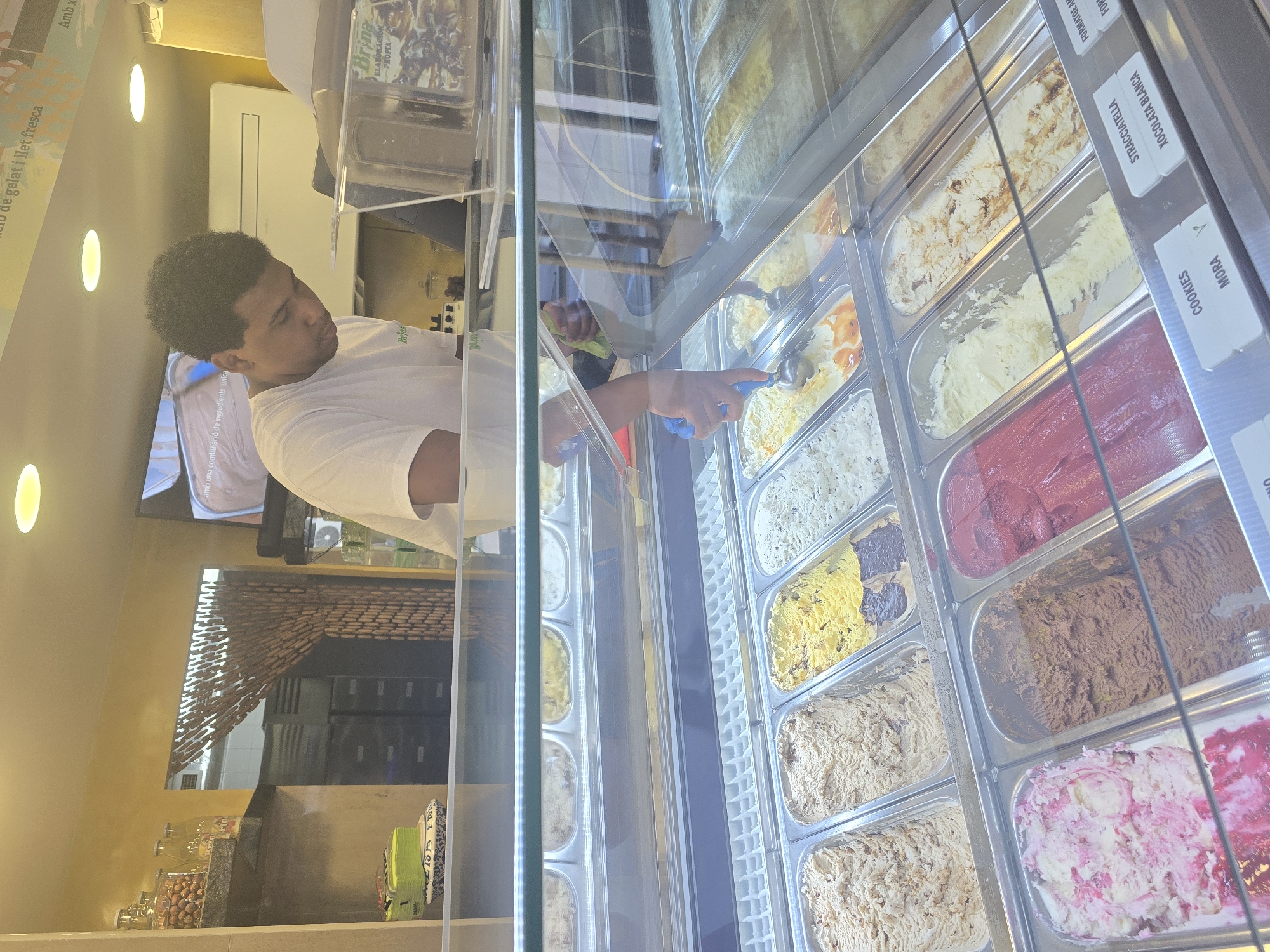 A man in a white t-shirt is scooping ice cream from a display case at an ice cream parlor, possibly in Cornellà de Llobregat, Spain, given the writing in Catalan. The case is full of different flavors, including cookies, mora (blackberry), stracciatella, and xocolata blanca (white chocolate). The parlor has a modern design, with mirrors, shelves, and jars of ingredients visible in the background.