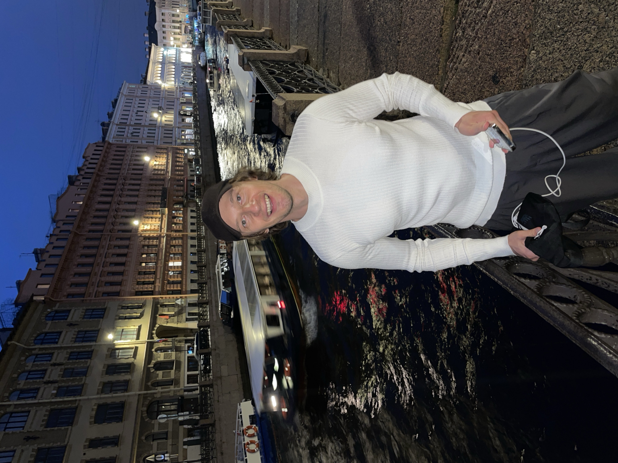An outdoor, eye-level medium shot captures a smiling man in a white, ribbed, long-sleeved sweater and grey pants standing on a cobblestone walkway next to a canal. The man wears a black baseball cap. His left hand holds a black cloth bag and a white cord, while his right hand holds a silver object, possibly a phone. Behind him, a narrow canal reflects the lights of buildings and boats. A boat with white seats is passing in the canal, and another boat is docked on the opposite bank. The buildings lining the canal are illuminated, and the sky is a deep blue, indicating twilight. The walkway is bordered by a decorative metal fence.