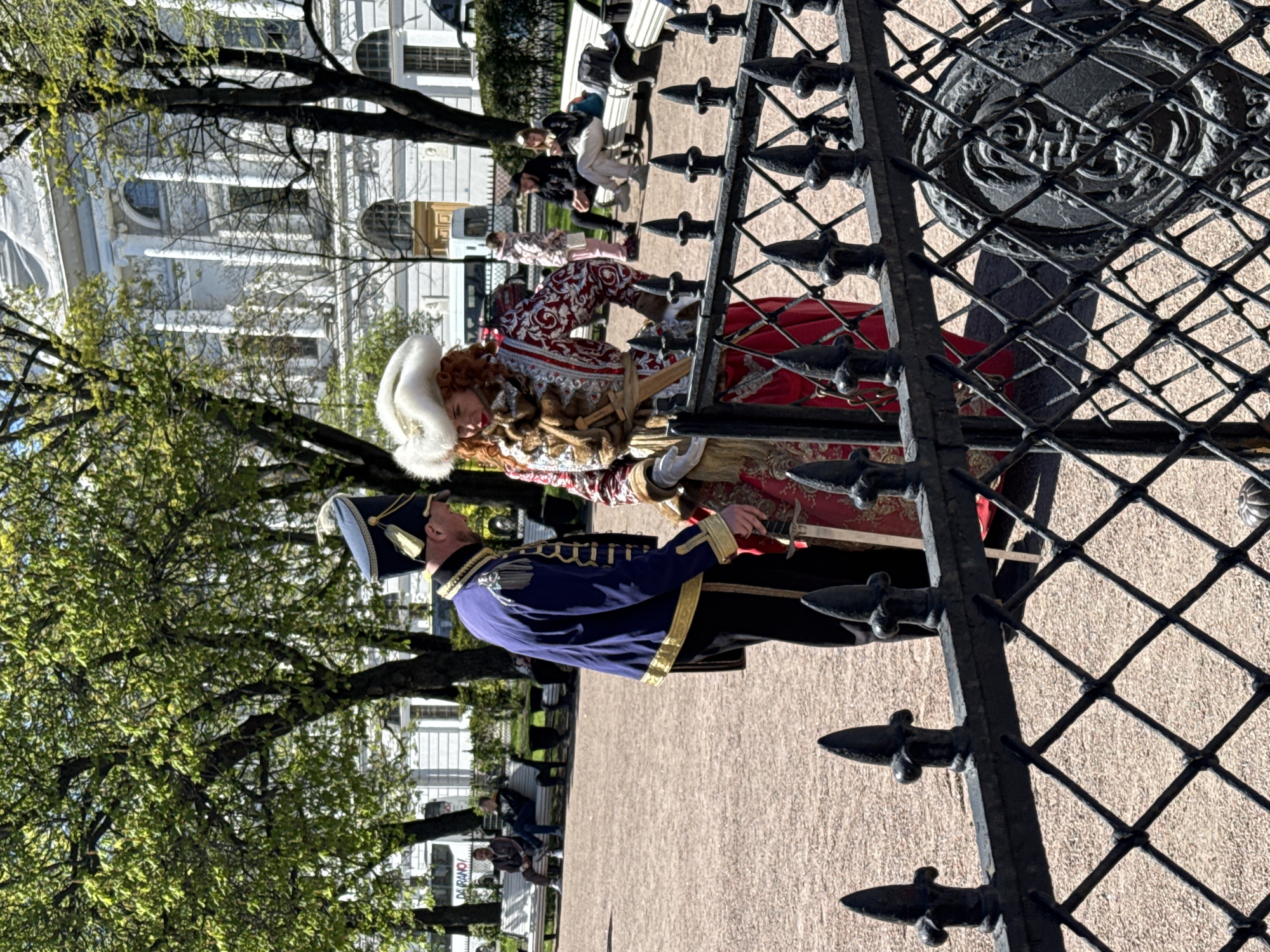 The image shows two people dressed in historical costumes, a man in a blue military uniform and a woman in a red and white dress and fur hat. They are standing in front of a metal gate with a decorative design. The background includes trees, a building with columns, and other people sitting on benches. The location appears to be a park or square in Saint Petersburg, Russia, near a building with classical architecture.