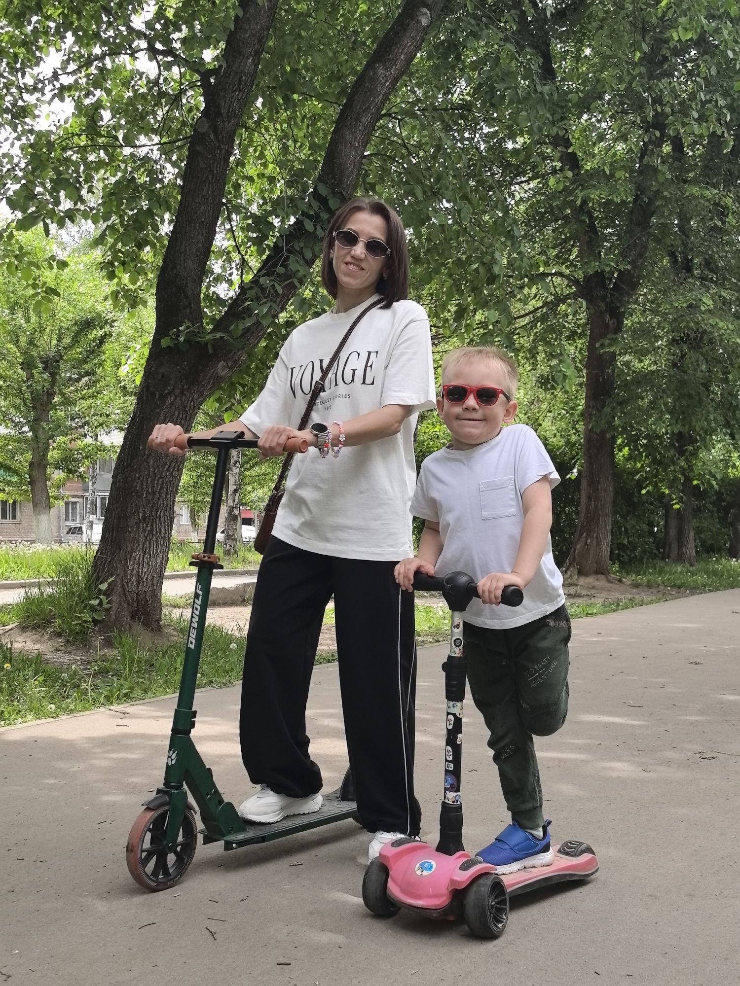 A mother and her young son pose outdoors with their scooters on a paved path in Perm, Russia. The woman, on the left, wears a white t-shirt with "VOYAGE" and smaller text "DELUXE STORIES 1970" printed on it, black wide-leg pants, and white sneakers. She has dark hair, oval sunglasses, and her wrist is adorned with a watch and beaded bracelets. She holds a dark green scooter, which has "DEWOLF" written on its frame.
To her right, the young boy, likely aged 3-5, wears a white t-shirt with a front pocket, dark green sweatpants with "NO PARTY ADVENTURE" visible, and blue sneakers. He sports bright red-framed sunglasses and holds a pink and black three-wheeled scooter. Both are smiling slightly towards the camera.
The setting appears to be a park or a tree-lined street, with lush green trees providing a dense canopy overhead. In the background to the left, glimpses of urban buildings can be seen. The lighting is soft, suggesting an overcast day during spring or summer.