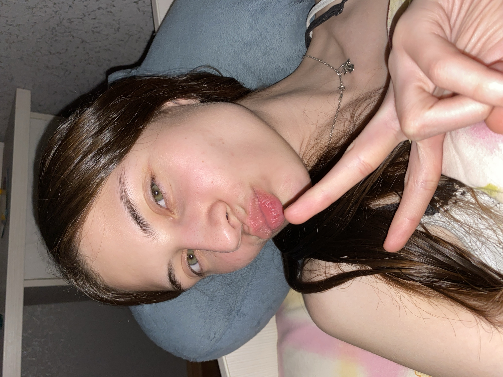 A young woman with long, dark hair is lying down, partially supported by a blue pillow. She is making a peace sign with her right hand, which is positioned close to her face. She is wearing a silver necklace and a patterned garment. The background includes a white wall and a pink and yellow patterned surface. There is no evidence in the image to suggest this image was taken in Votkinsk, Russia.
