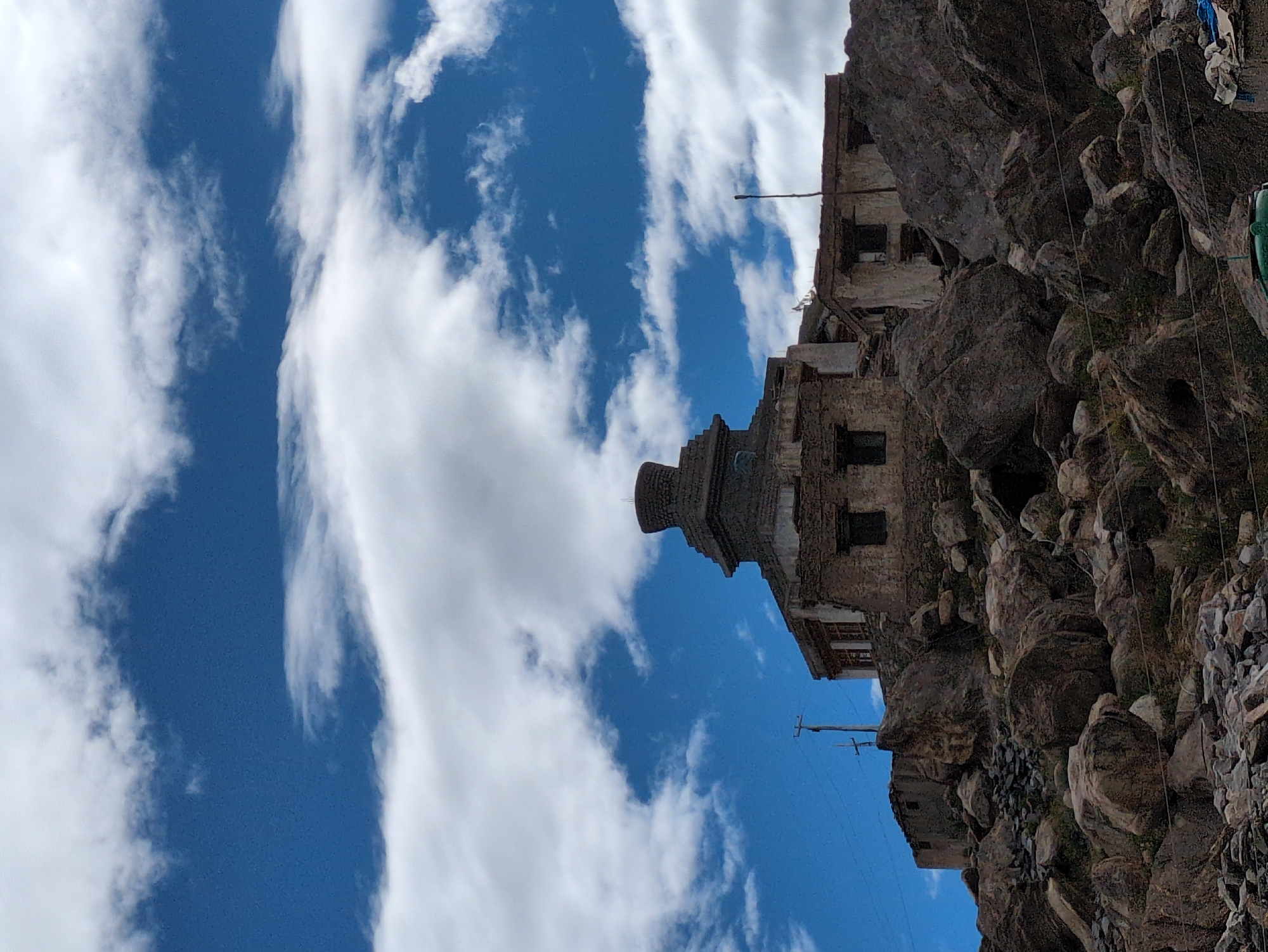 An upward-angle, medium shot captures a Buddhist stupa atop a rocky, barren hillside in Padum, India. The stupa, constructed of dark stone, is segmented into a square base, a circular drum, and a cone-shaped finial. The surrounding buildings, also of stone, are weathered and appear partially ruined, with visible windows and doorways. The hillside is densely covered with large, rough-hewn rocks. In the background, a bright blue sky is filled with scattered white clouds. Telephone poles and wires are visible on the left side of the frame, indicating the presence of infrastructure. The overall scene depicts a remote and rugged landscape with ancient architecture.