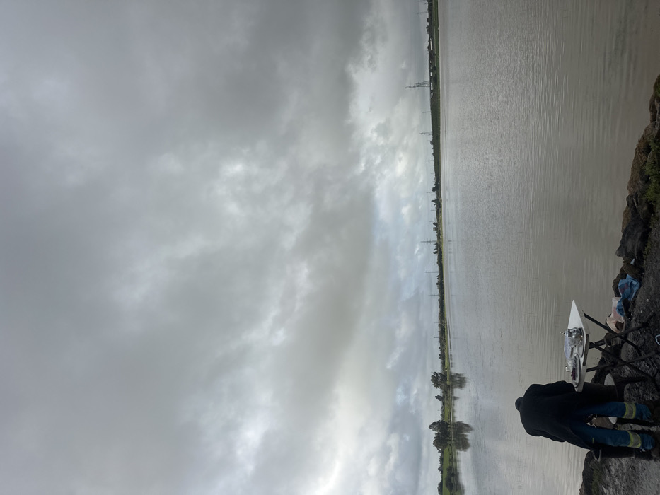 The image depicts an outdoor scene in Oumazza, Morocco, under a heavily overcast sky dominated by dense grey clouds. A single individual, observed from behind, is bent forward, facing a small white rectangular folding table positioned on a muddy bank at the edge of a large, calm body of water. The person wears a dark, long overcoat and blue trousers with prominent reflective yellow-green stripes near the ankles, along with dark boots. On the table, a silver teapot, at least two clear glass cups, and a dark rectangular tray are visible. The immediate ground around the table is muddy and dark, with scattered rocks and what appears to be a crumpled blue plastic bag. The expansive body of water is a dull grey-brown, exhibiting minimal ripples and reflecting the overcast sky and distant landscape. Along the distant left shoreline, green fields and a cluster of trees are visible, with their reflections mirrored in the water. Stretching across the horizon on the right side of the image is a line of electricity pylons, with indistinct structural elements visible further in the distance.