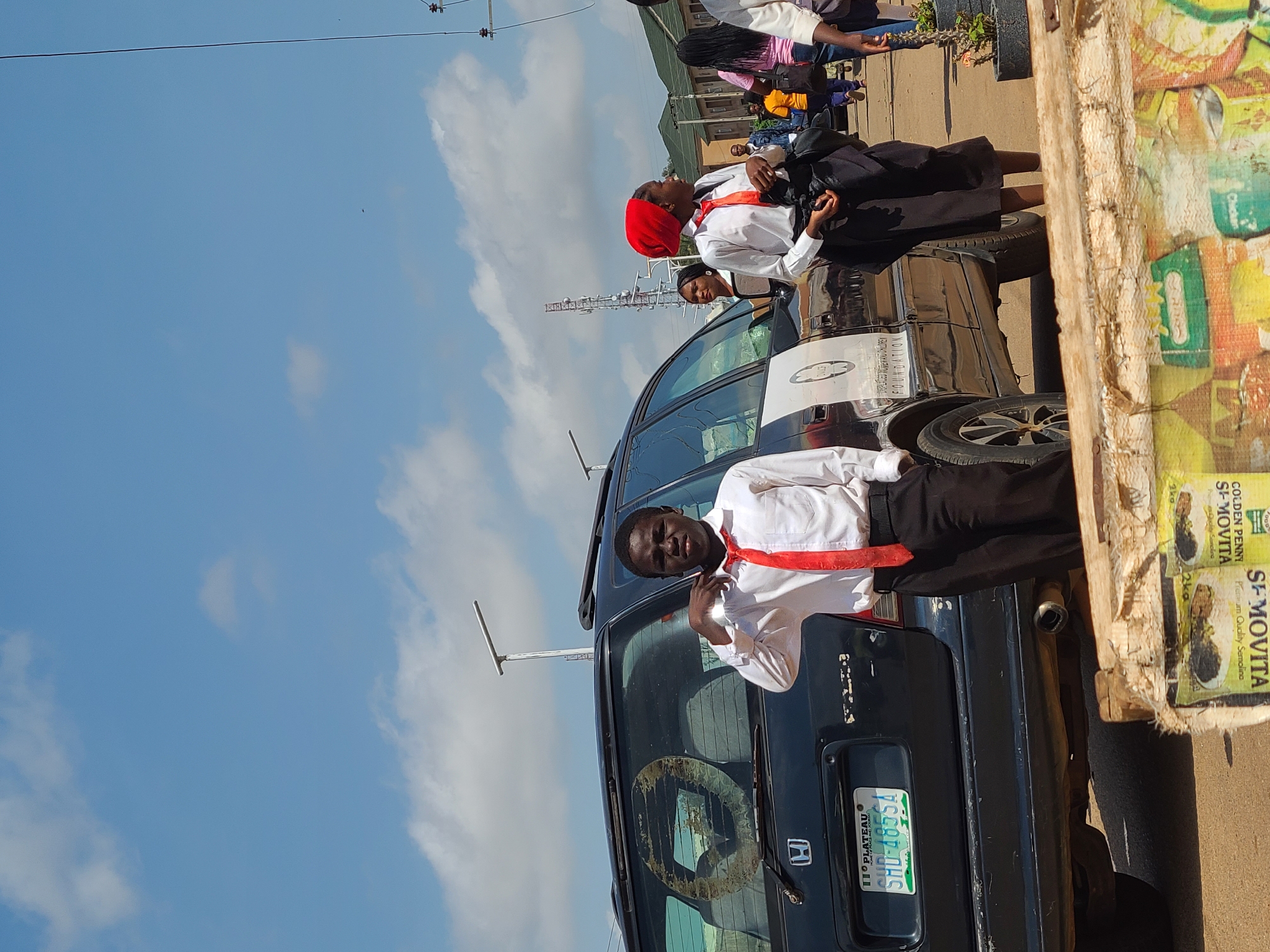 In Jos, Nigeria, a young boy in a white shirt and red tie stands next to a black SUV. Behind him, a young girl in a white shirt, red headscarf, and black skirt also stands near the vehicle. The SUV has a license plate that reads "PLATEAU SHD 4855A" and has text on its side. In the background, other people walk by, and a telecommunications tower is visible. The sky is blue with scattered clouds. A wooden structure with what appears to be bags of product is in the foreground.