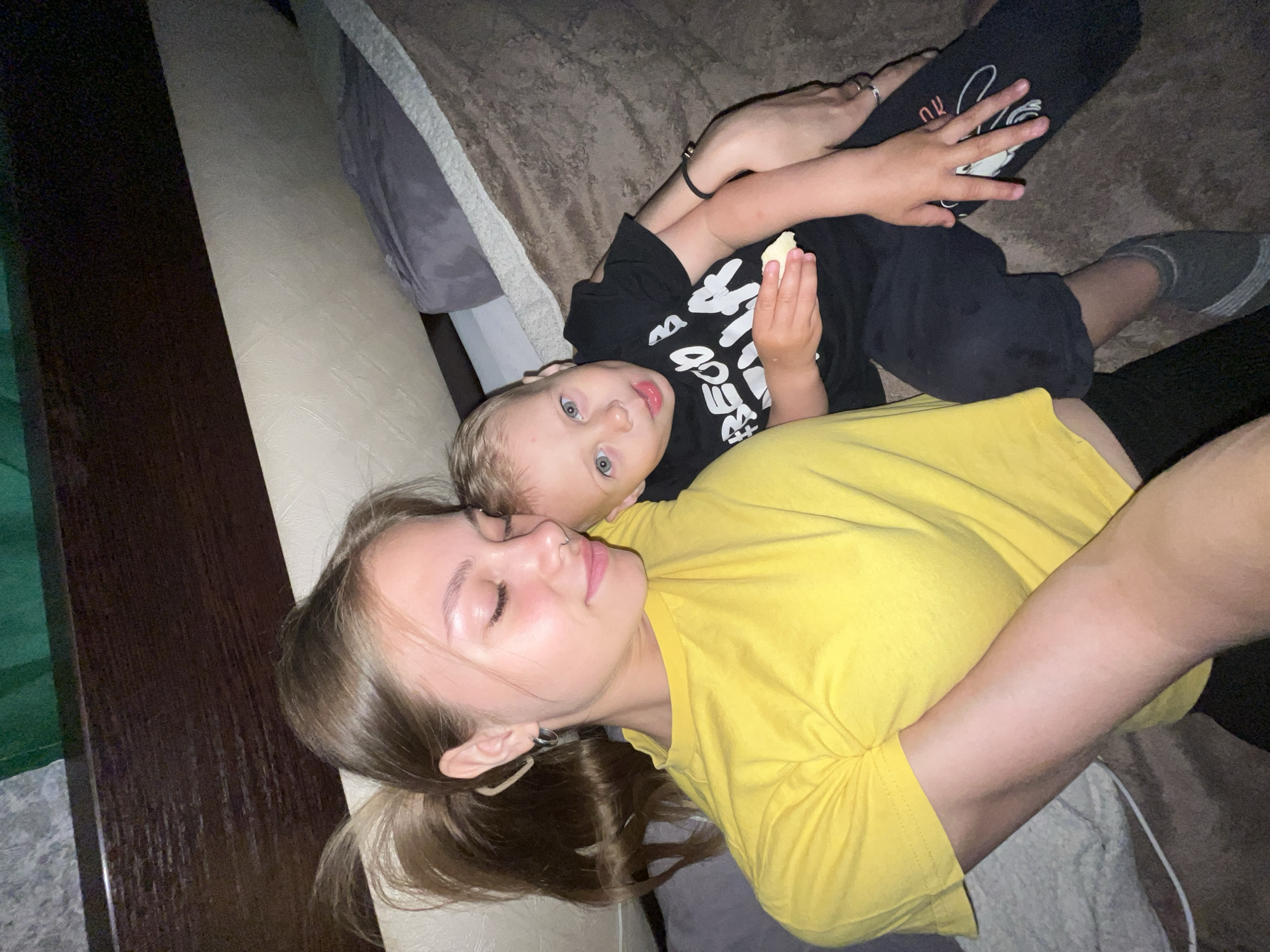 A woman and a child are lying on a bed. The woman has fair skin, light brown hair in a ponytail, a nose ring, and is wearing a yellow t-shirt. The child has light skin, blonde hair, blue eyes, and is wearing a black t-shirt with white writing. The child is holding a piece of food. The bed has a brown headboard and a gray blanket. The photo was taken indoors. The location is Фоки, Russia.