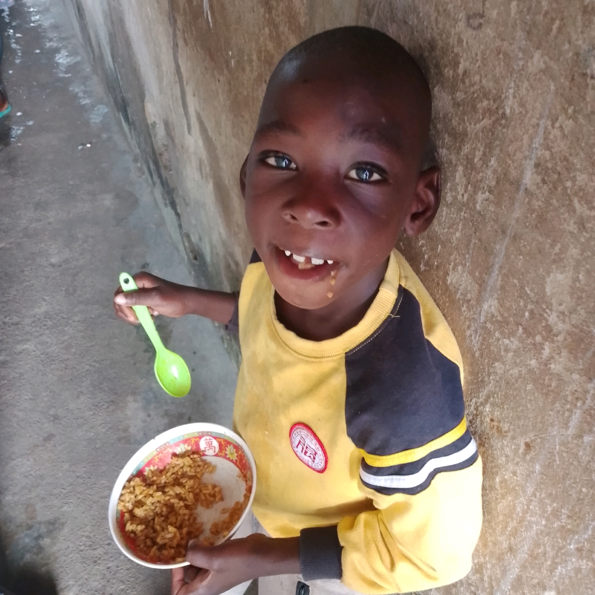In Ado, Nigeria, a young boy with dark skin and short hair is shown looking towards the camera. He is wearing a yellow and black long-sleeved shirt with yellow trim on the collar and sleeves. In his left hand, he holds a white bowl filled with rice, and in his right hand, he holds a green spoon. The boy's mouth is open, with some food visible on his teeth and a drip of food extending from his chin. He is standing against a textured, light brown wall. The ground in front of him appears to be concrete and slightly wet.