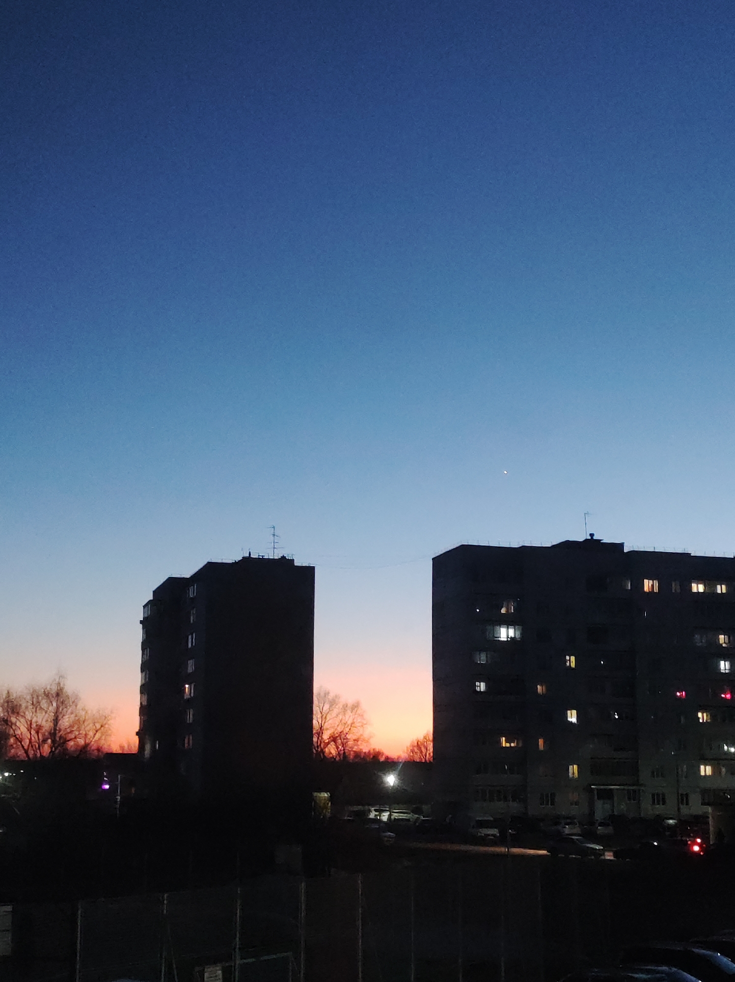 The image shows two apartment buildings in Chaikovsky, Russia at dusk. The sky transitions from blue to orange. Lights are on in some of the apartments in the building on the right. Trees and cars are visible in the foreground. A chain link fence is in the lower part of the image. An antenna is on top of each building. There's a single bright spot in the sky above the building on the right.