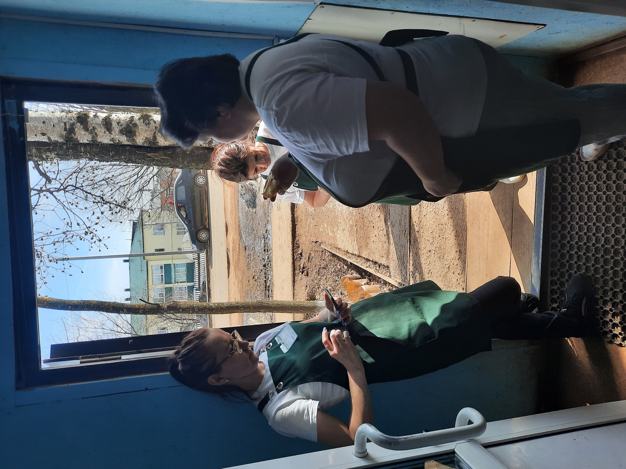 The photo, apparently taken from inside a building in Krasnogorskoye, Russia, shows three people. One woman, wearing a white shirt and a green pinafore, is sitting at a window, holding a mobile phone. Another woman, wearing a white shirt and a green apron, is standing on the outside of the building, presumably talking to the woman at the window. A third woman, also wearing a white shirt and green apron, stands behind the second woman, holding an item in her hand. Outside the window, there is a view of a house, a car, trees without leaves, and a dirt ground.