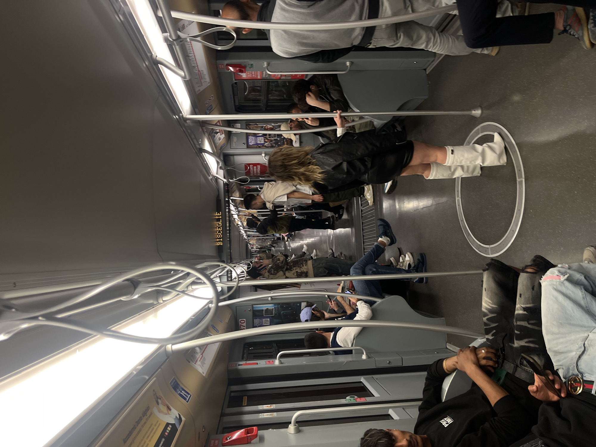 The image depicts the interior of a subway car, likely in Milan, Italy, as the station name "Bisceglie" is visible. The car is occupied by several people of various ages and attire, some standing and some seated. A woman in a black leather jacket and white boots stands near a circular floor marker. A man wearing a black hoodie lies back across seats on the right. Other passengers are visible in the background, some holding onto handrails or seated in the grey seats. The interior is lit by overhead fluorescent lights and features numerous handrails. Advertisements and route maps can be seen along the walls.