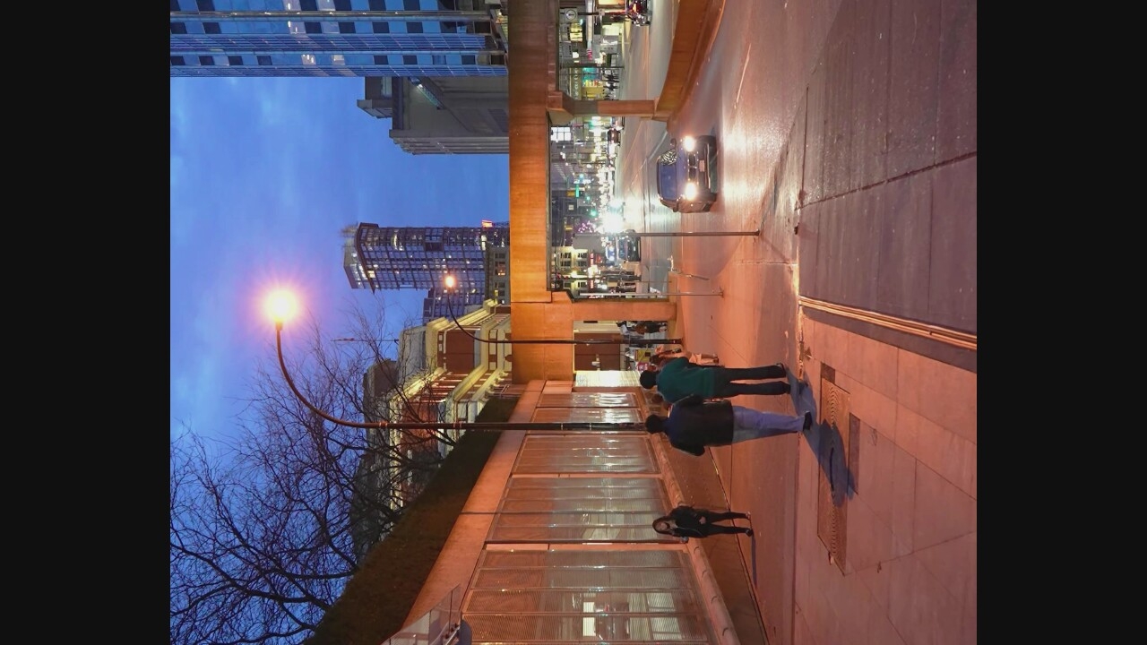 The image captures a street scene at dusk in Vancouver, Canada. The perspective is from a sidewalk looking down a city street. Tall buildings are visible in the background, including a modern skyscraper with a lit top. A concrete overpass spans the street in the mid-ground. Several people are walking on the sidewalk in the foreground and mid-ground. One person is standing alone against a textured wall. Cars are driving on the street, with headlights illuminating the wet pavement. A streetlamp is visible in the upper left portion of the image, casting a warm glow. The sky is a deep blue, indicating the transition from day to night. The overall impression is of a busy urban environment at the end of the day.
