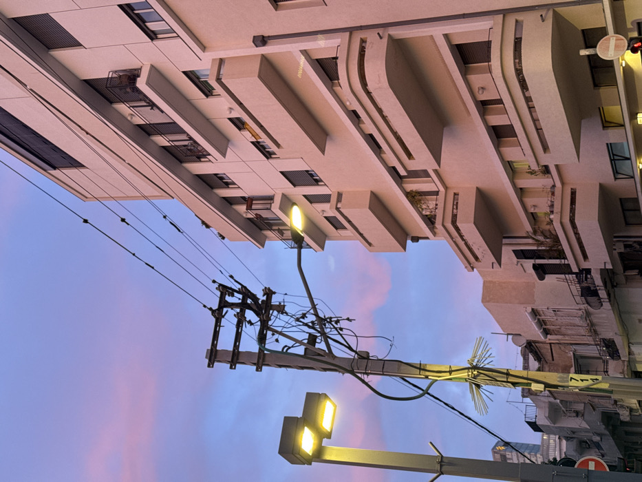 An upward view captures architecture against a pastel sky. On the left, a modern building with balconies and windows angles across the frame. Utility poles intersect the scene, tangled with wires and lit by streetlights. Other buildings stand on the right, their architecture less distinct. The sky transitions from light blue to pink, adding a soft contrast to the angular structures.