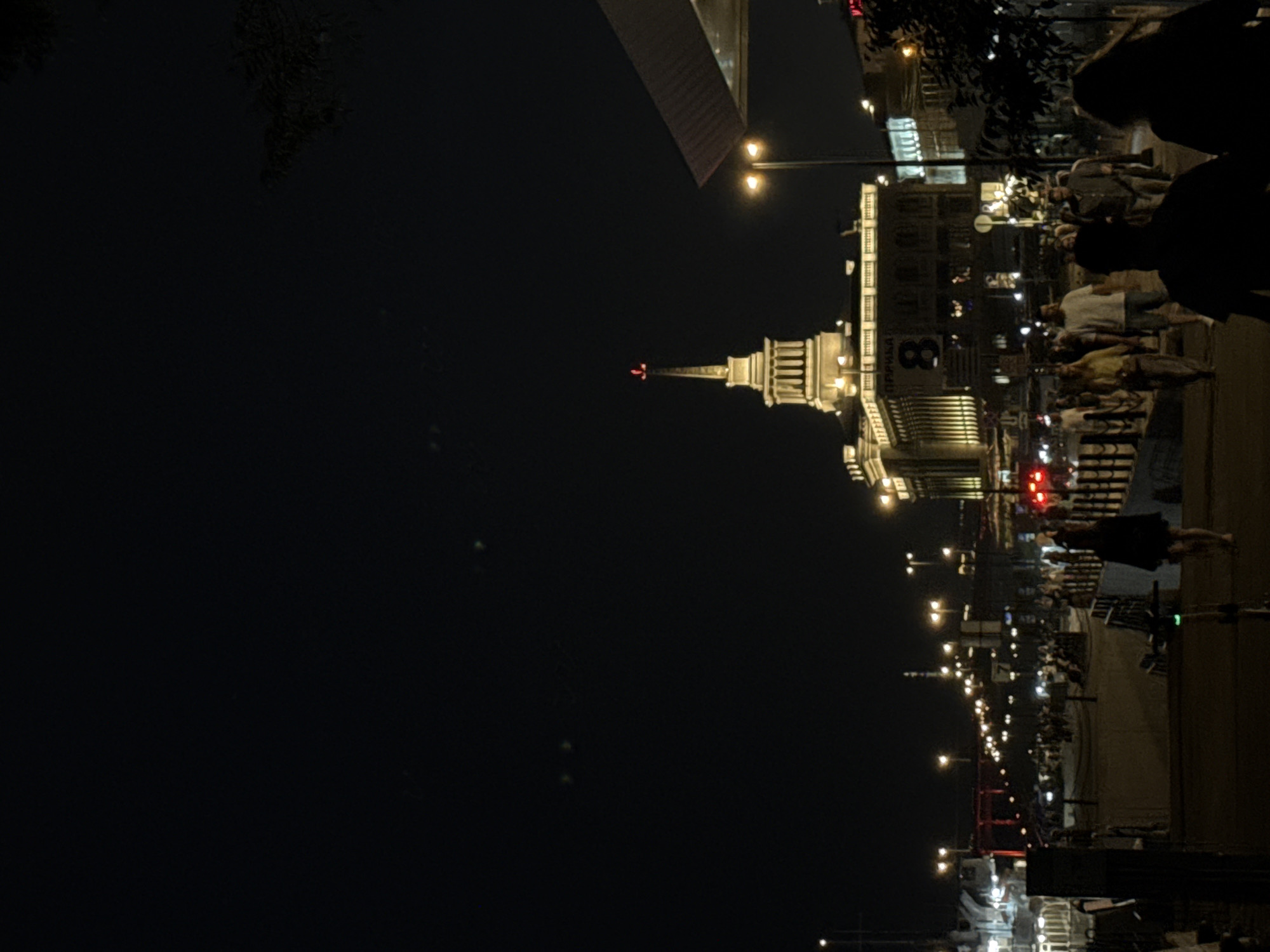 The image depicts a nighttime scene along the embankment in Krasnoyarsk, Russia. A prominent building with a spire is illuminated, showcasing its architectural details. Several streetlights are visible, casting a warm glow on the area. A crowd of people are walking along the embankment, some appearing to be engaged in conversation. A red bridge is visible in the distance, further contributing to the cityscape. The overall atmosphere is one of a lively urban environment at night.