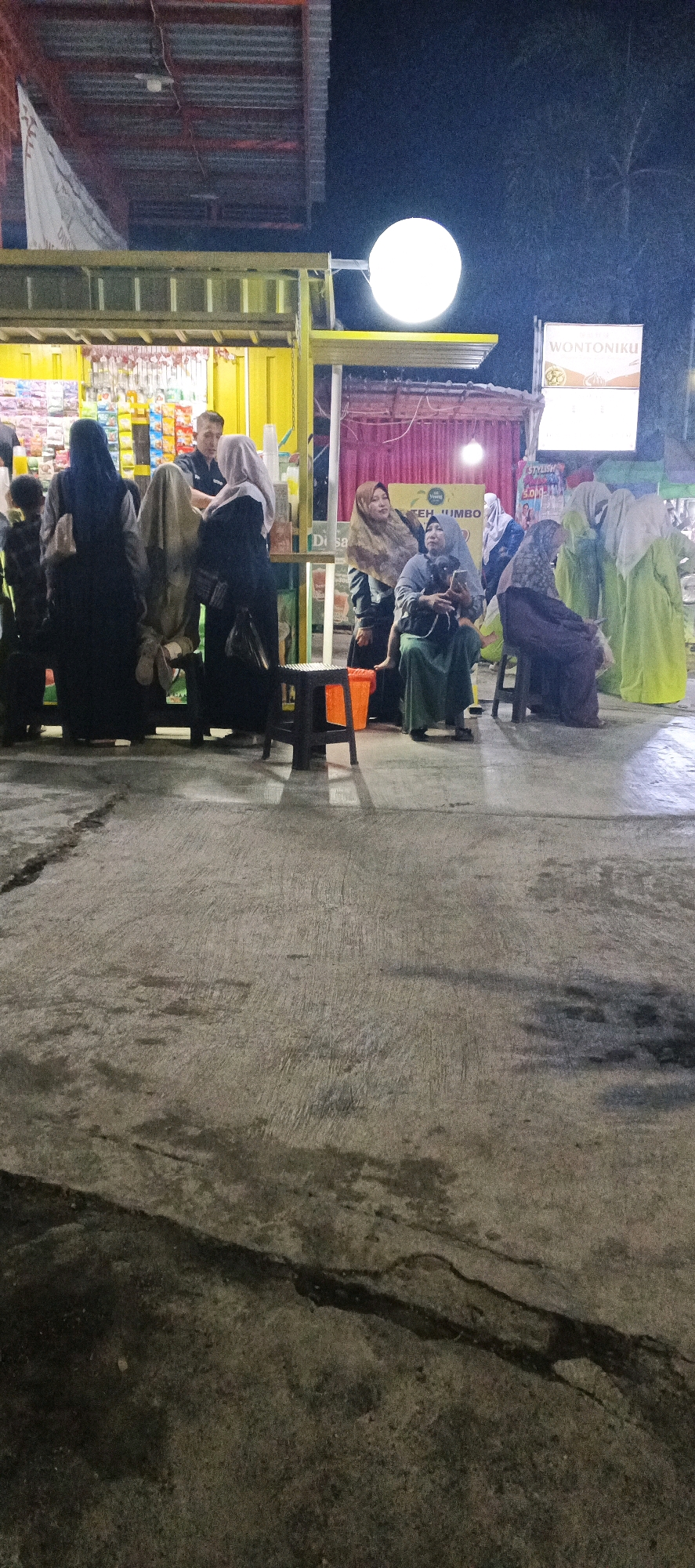 In Sungelebak, Indonesia, at night, a group of people are gathered outside a brightly lit food stall. The stall, with its yellow facade, displays various snacks and drinks behind a counter. Several individuals, predominantly women wearing hijabs and long garments in black, beige, and light green, are standing or seated on stools. One woman in the center, wearing a grey hijab and dress, holds a small black dog. A man in a dark shirt stands behind the counter, interacting with customers. The ground in the foreground is paved with concrete and appears wet, with some cracks and dark stains. Overhead, a large, round, white light illuminates the scene, and a sign reading "WONTONIKU" is visible to the right. The overall atmosphere suggests a casual evening gathering at a local eatery.