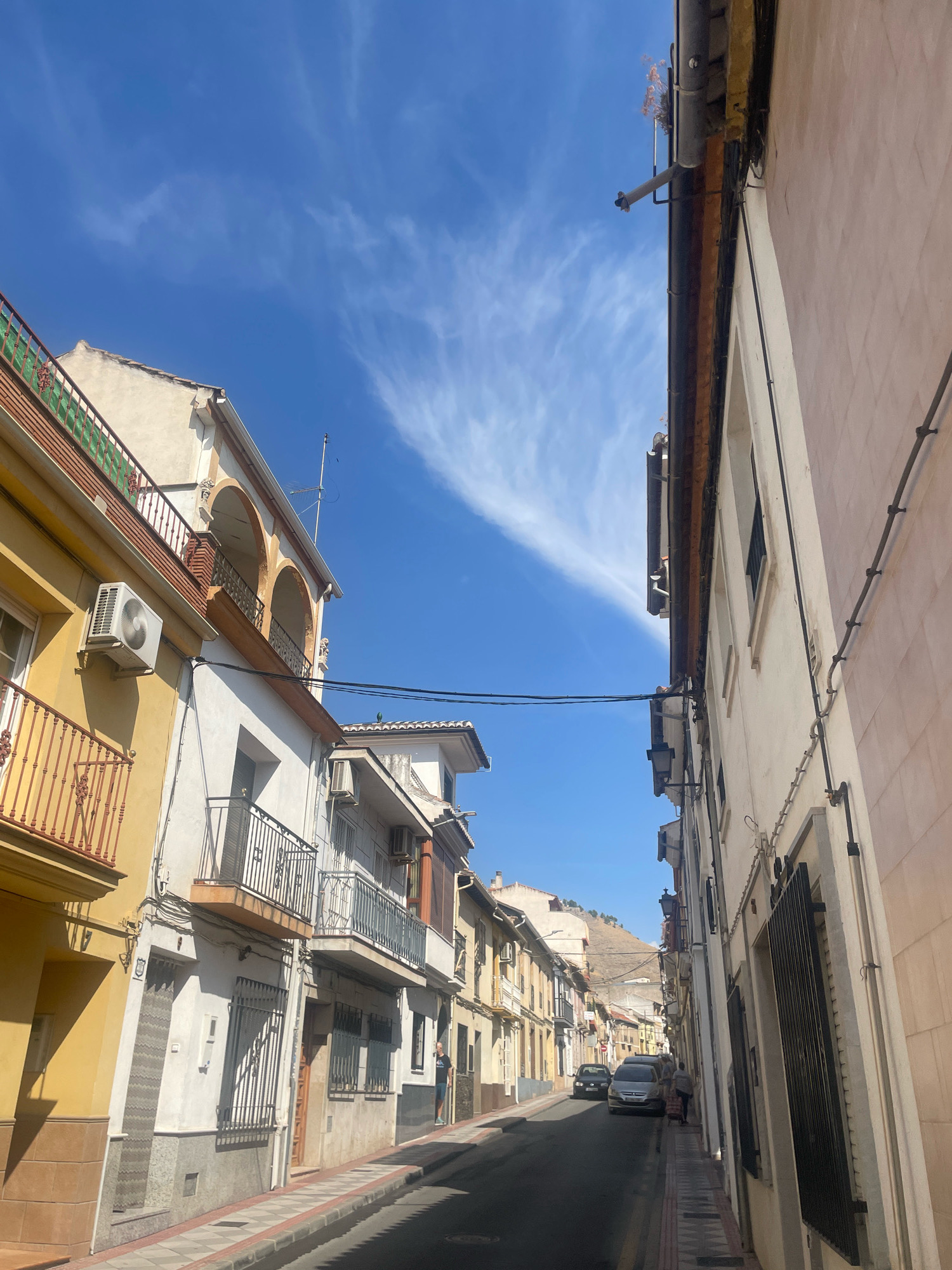 The image shows a street scene in Atarfe, Spain, on a sunny day with a bright blue sky and wispy white clouds. The viewpoint is from a street looking down a narrow, winding road lined with multi-story buildings. The architecture appears to be residential, with balconies, windows, and air conditioning units visible on the building facades.
On the left side of the street, a yellow and white building features a red-railed balcony on the second floor and an air conditioning unit mounted on the wall. Further down, a white building has a similar balcony and architectural details.
On the right side of the street, a pale pink building with a dark-tiled roof runs along the side. Electrical wires and conduits are attached to the exterior wall.
Several cars are parked or moving along the street. One silver car is parked on the right side, and a dark car is further down the street. A person wearing a dark t-shirt and blue shorts is walking on the sidewalk on the right side of the street, pushing a cart. Another person is partially visible in the distance, standing near the silver car.
The street surface is asphalt, with a sidewalk on the right made of paving stones. The ground has a gradient, suggesting the street may be on a slope. In the background, a hillside is visible, with some buildings scattered on it. The overall impression is of a typical Mediterranean town street.